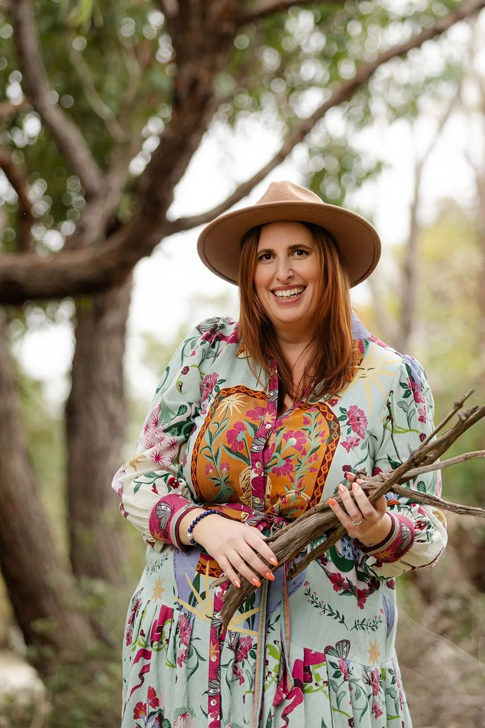 Smiling Nicole Andrews with red hair wearing a wide-brimmed brown hat and a colourful dress, standing outdoors amidst trees, holding sticks for camping.