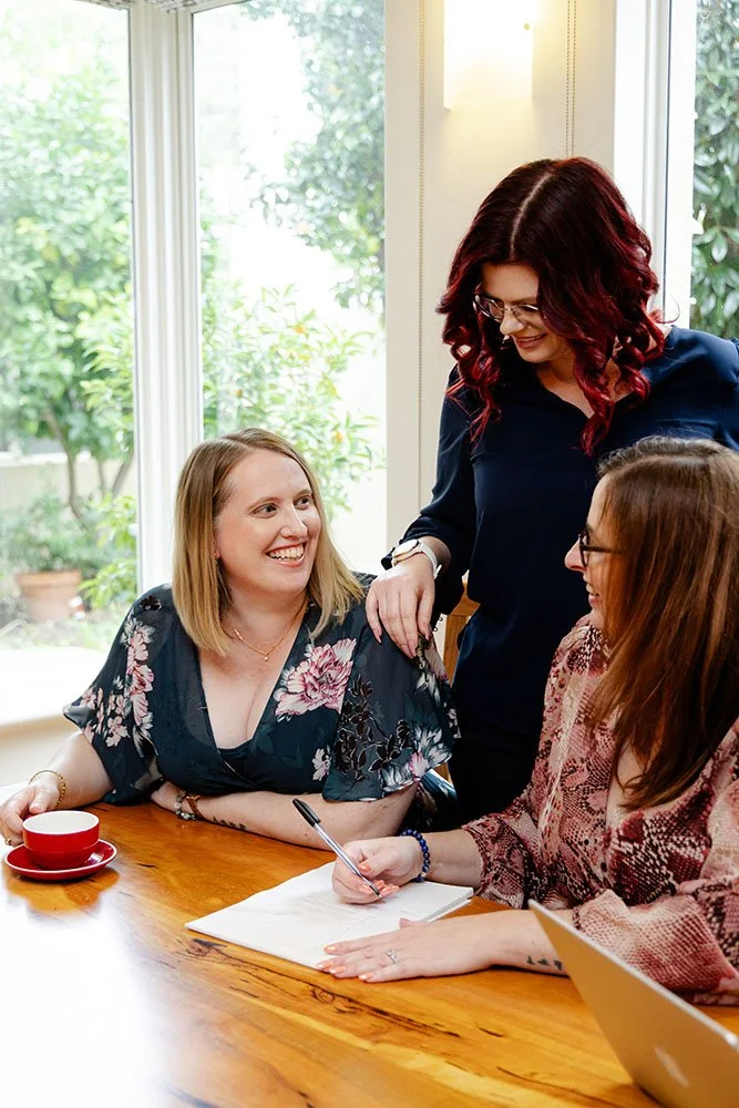 Three women of the Firefly Financil team having a conversation at a wooden table in a bright room with a large window showing greenery outside. There are notebooks, a pen, a laptop, and a red cup on the table.