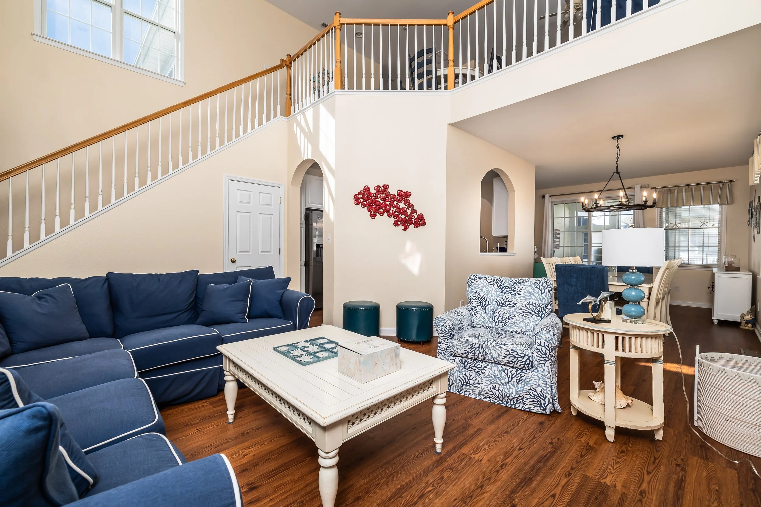 Living room with blue couches, patterned armchair, white coffee table, dark wood flooring, second-floor balcony, chandelier, and windows.