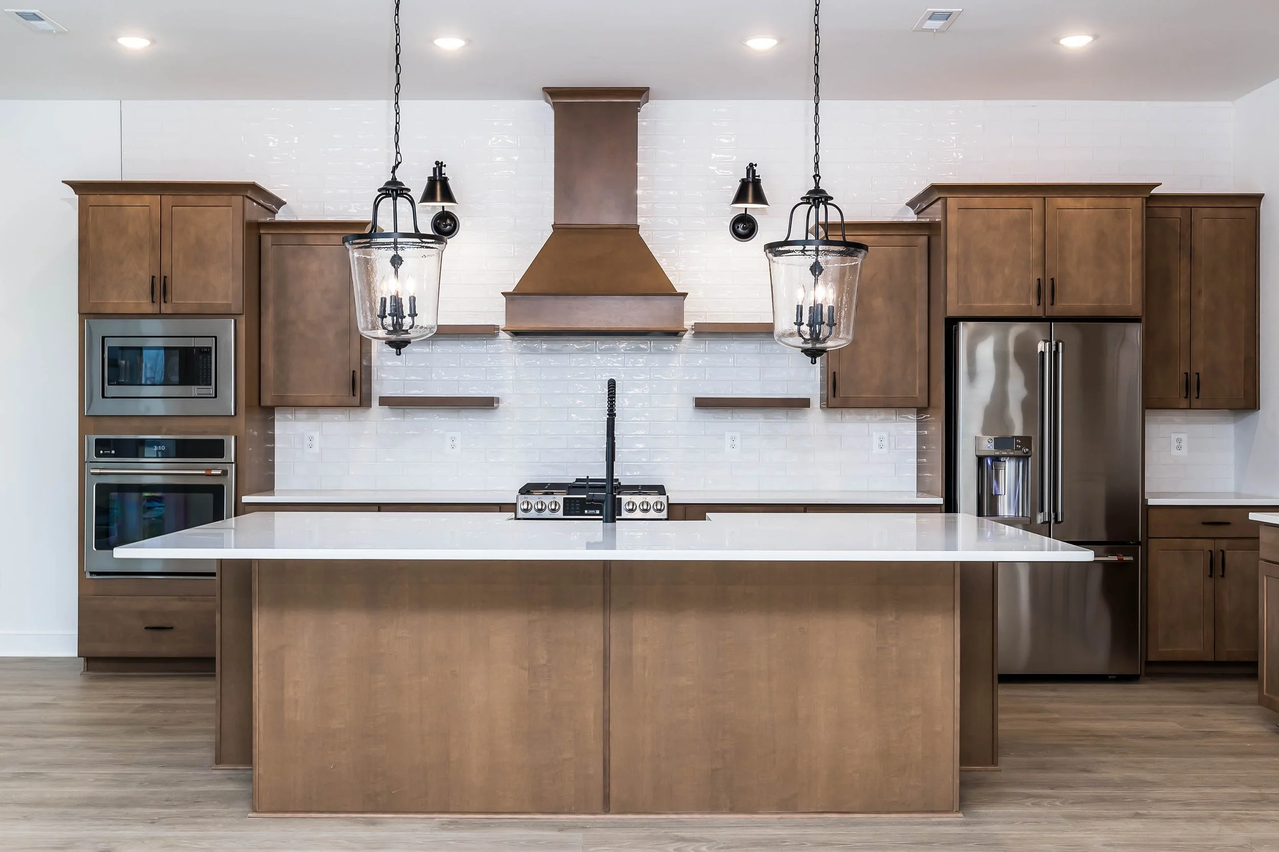 Modern kitchen with brown wooden cabinets, white subway tile backsplash, and stainless steel appliances including a microwave, oven, and refrigerator. A large white island with a black sink is in the foreground. Two black pendant lights with glass co