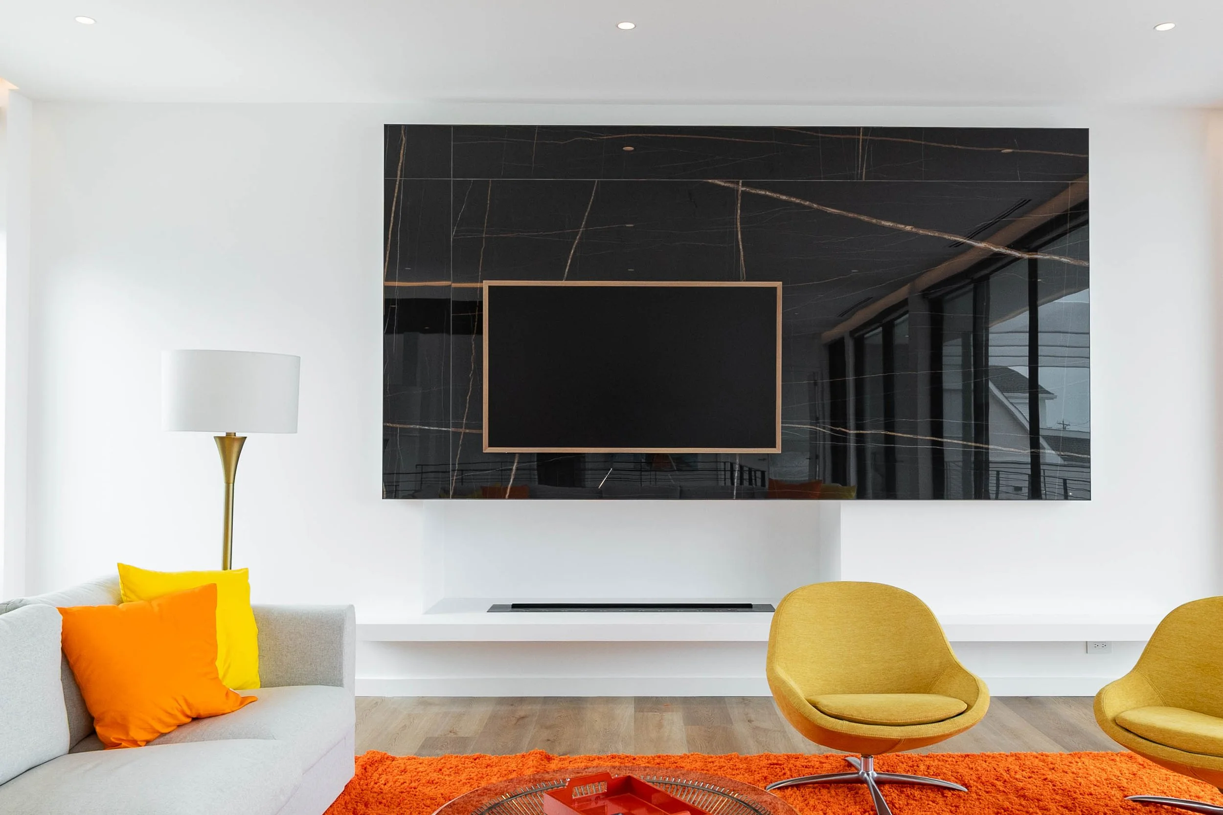 A modern living room with a white sofa adorned with orange and yellow pillows, a yellow accent chair, a tall white and gold floor lamp, a black marble wall with gold veining, a large flat-screen TV, and a bright orange shag rug.