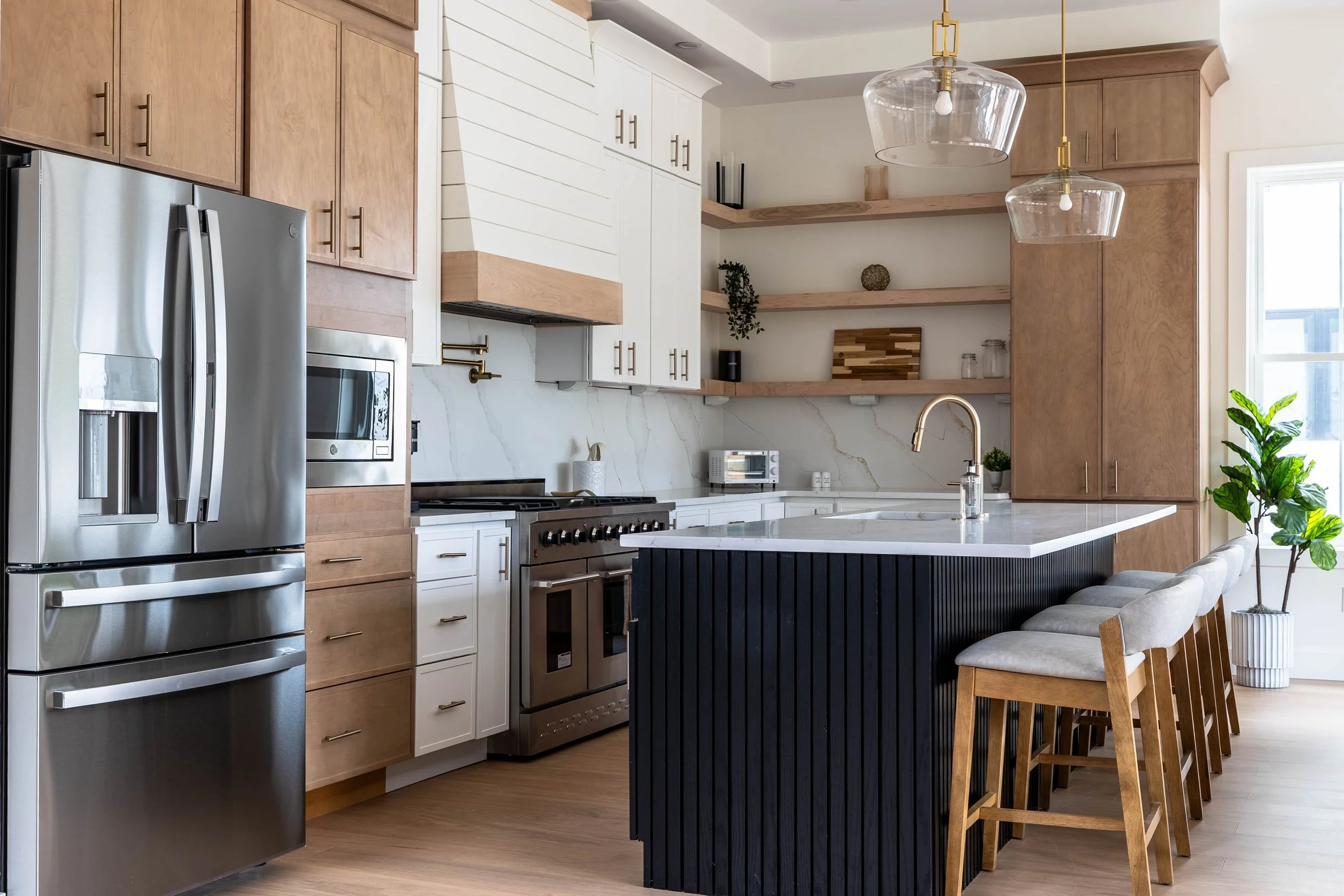 Modern kitchen with stainless steel refrigerator, white and wood cabinets, open shelves, marble backsplash, black kitchen island, and gray cushioned chairs at the island, with a large window and potted plant nearby.