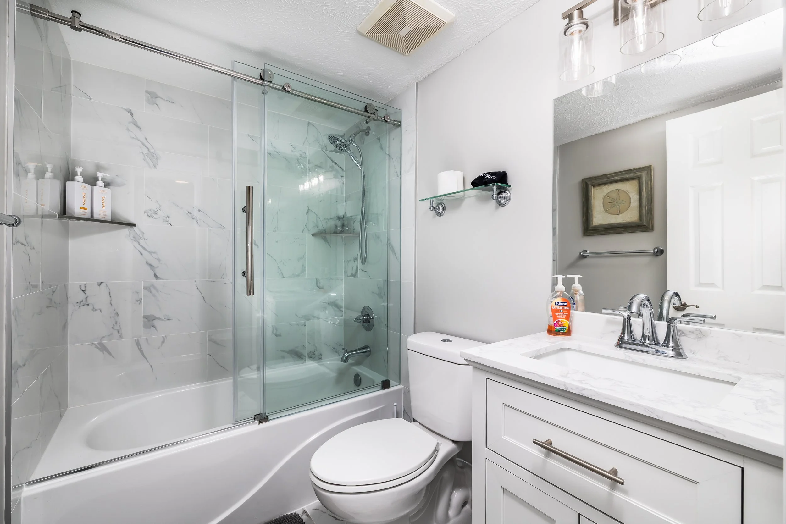 Modern bathroom with a glass enclosed shower, white bathtub, toilet, and a white vanity with a marble countertop. Mounted glass shelf with toiletries, bathroom mirror, towel rack, and framed artwork on the wall.