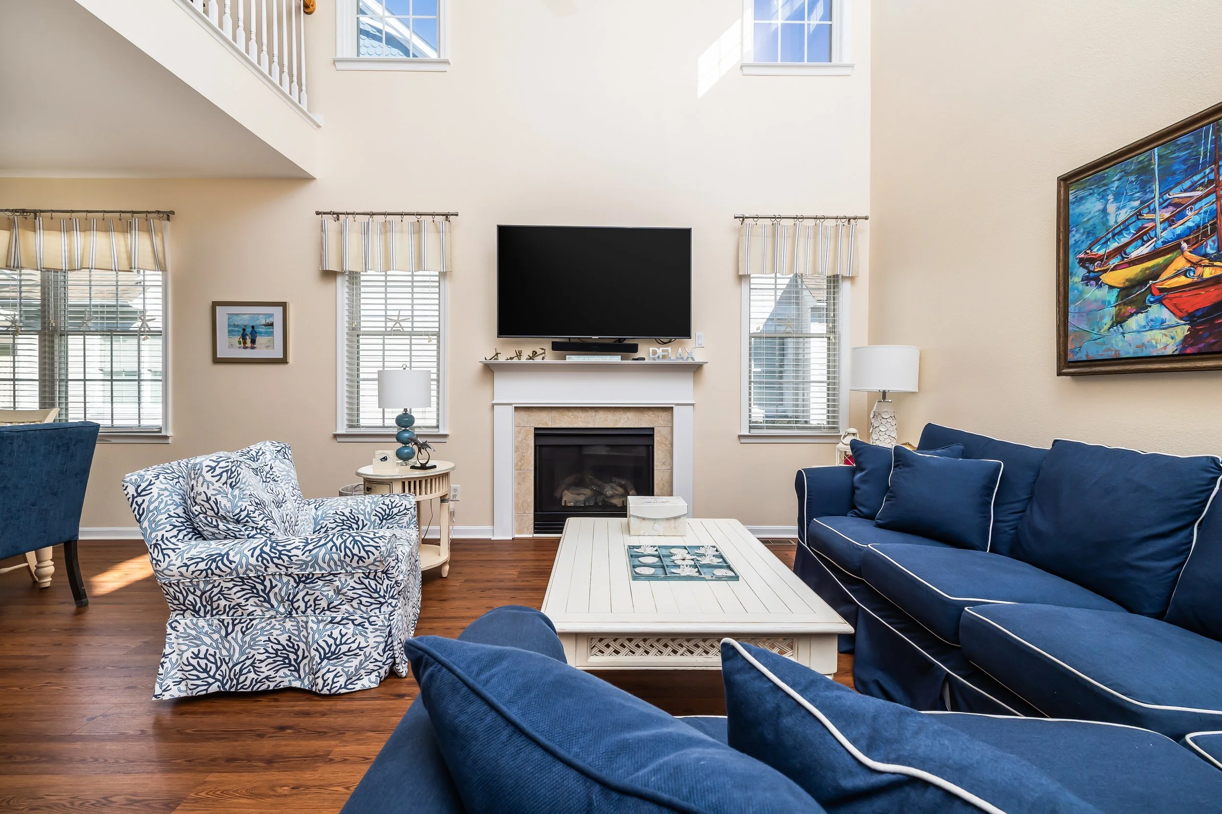 Living room with blue sofas, patterned armchair, white coffee table, fireplace, wall-mounted flat-screen TV, and windows with white blinds and curtains.