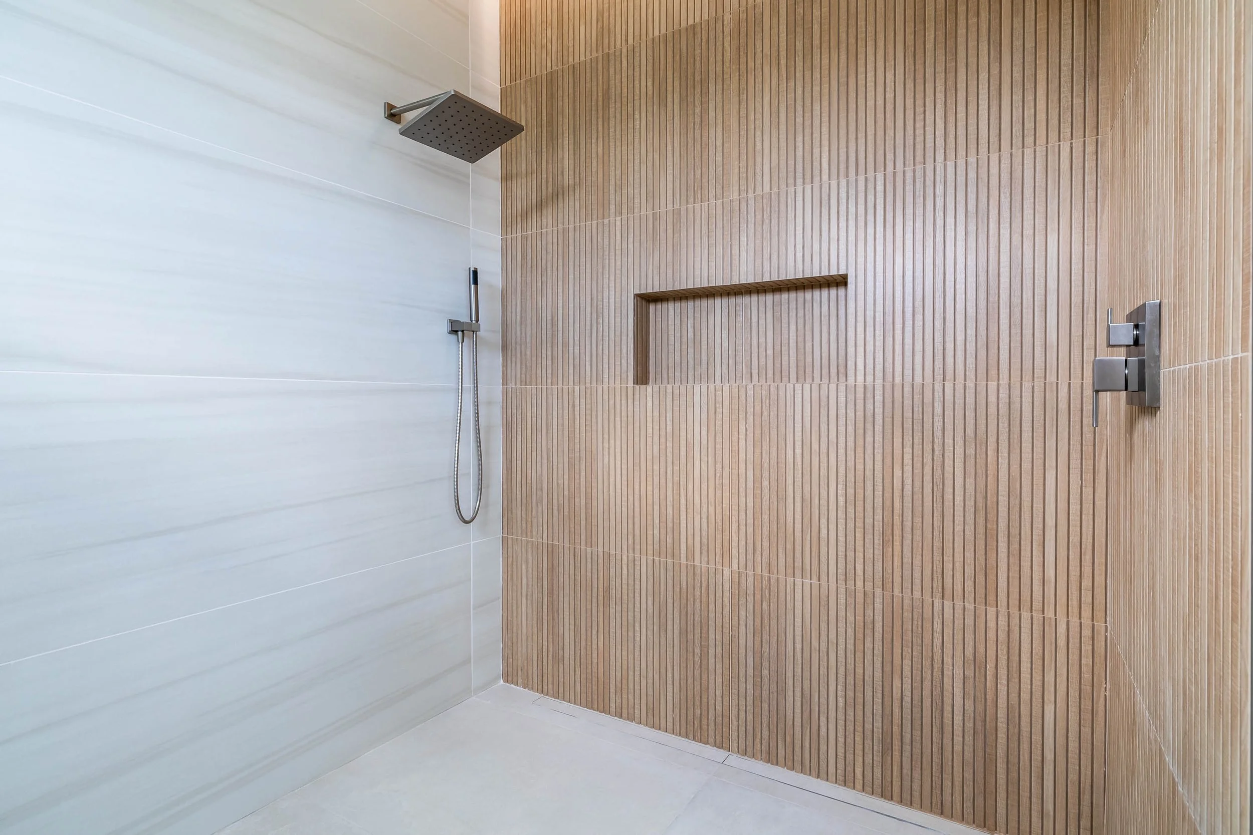 A modern shower area with a large square rainfall showerhead attached to the ceiling, a handheld shower on the wall, and a built-in wooden shelf, featuring light-colored wall tiles and wooden paneling.