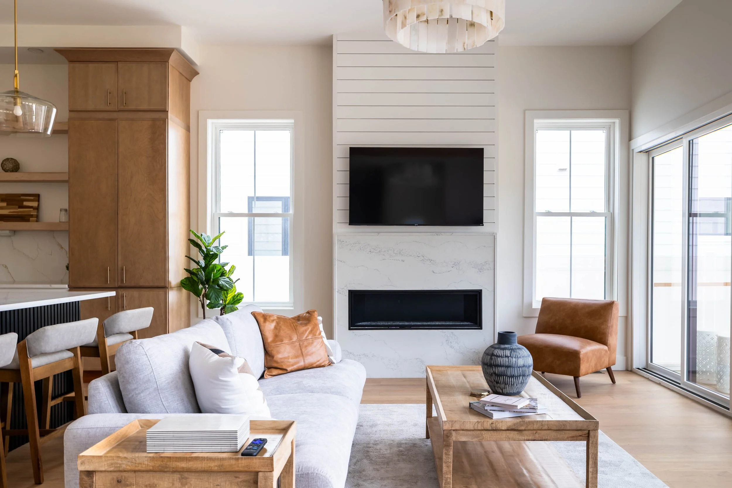 Modern living room with white sofa, brown leather chair, wooden coffee table, fireplace, flat-screen TV, large windows, and decorative vase.