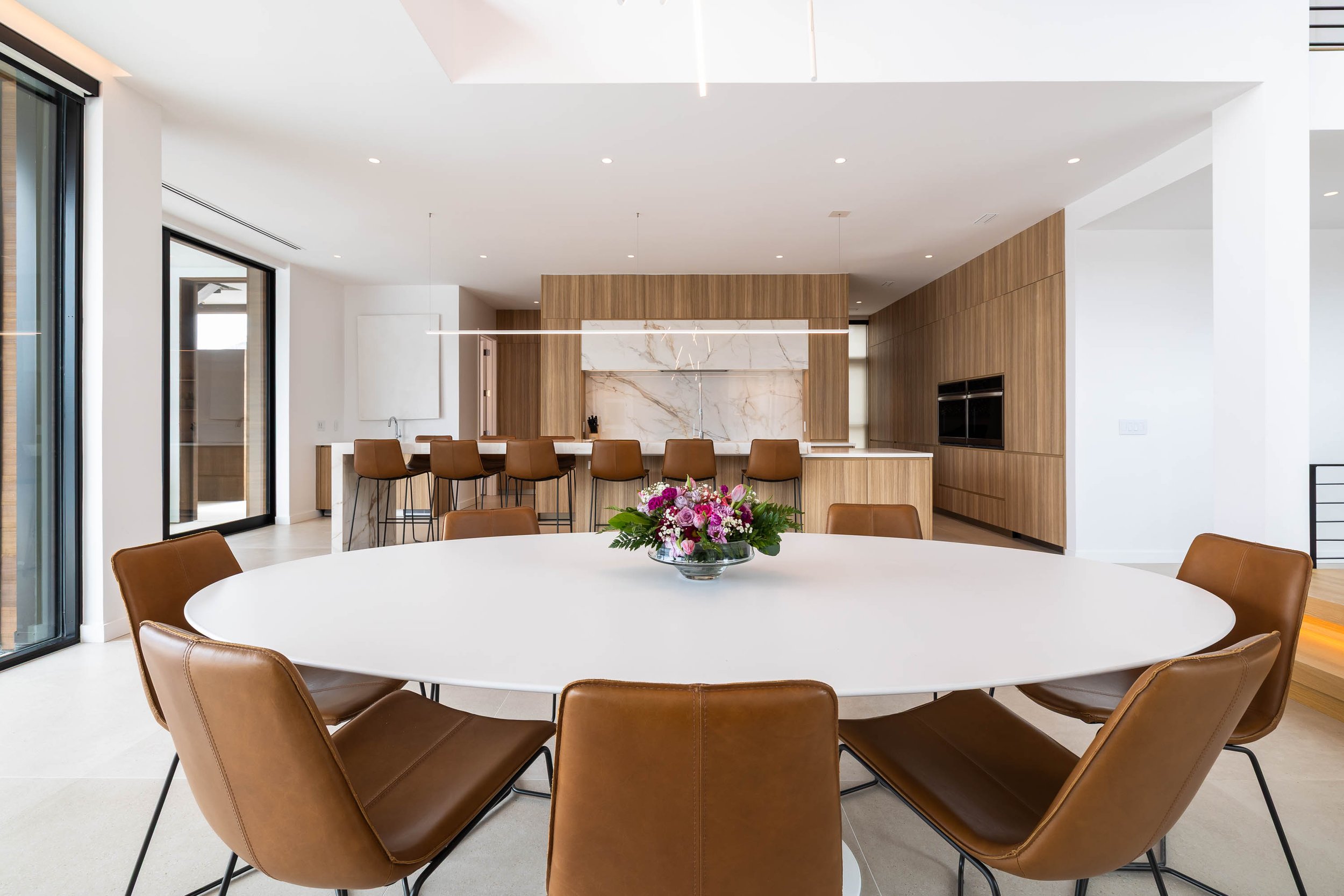 Modern dining area with a large round white table surrounded by brown leather chairs, a colorful flower arrangement in a glass bowl at the center of the table, and a backdrop of wooden wall panels and built-in cabinets.