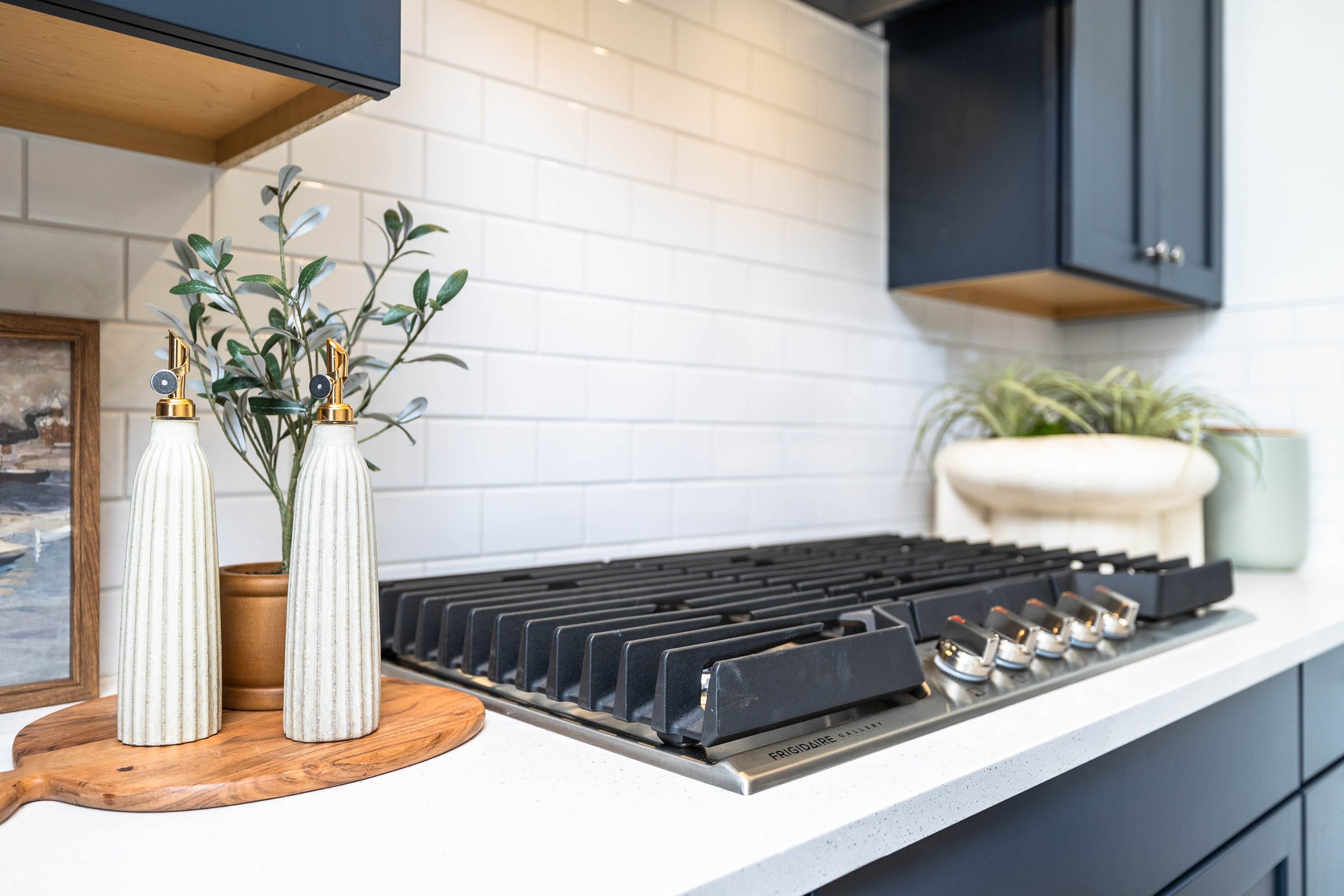 Kitchen with white subway tile backsplash, black stove with silver knobs, and dark cabinets. On the counter, there are two tall vases with green plants, placed on a wooden tray along with a framed photo.