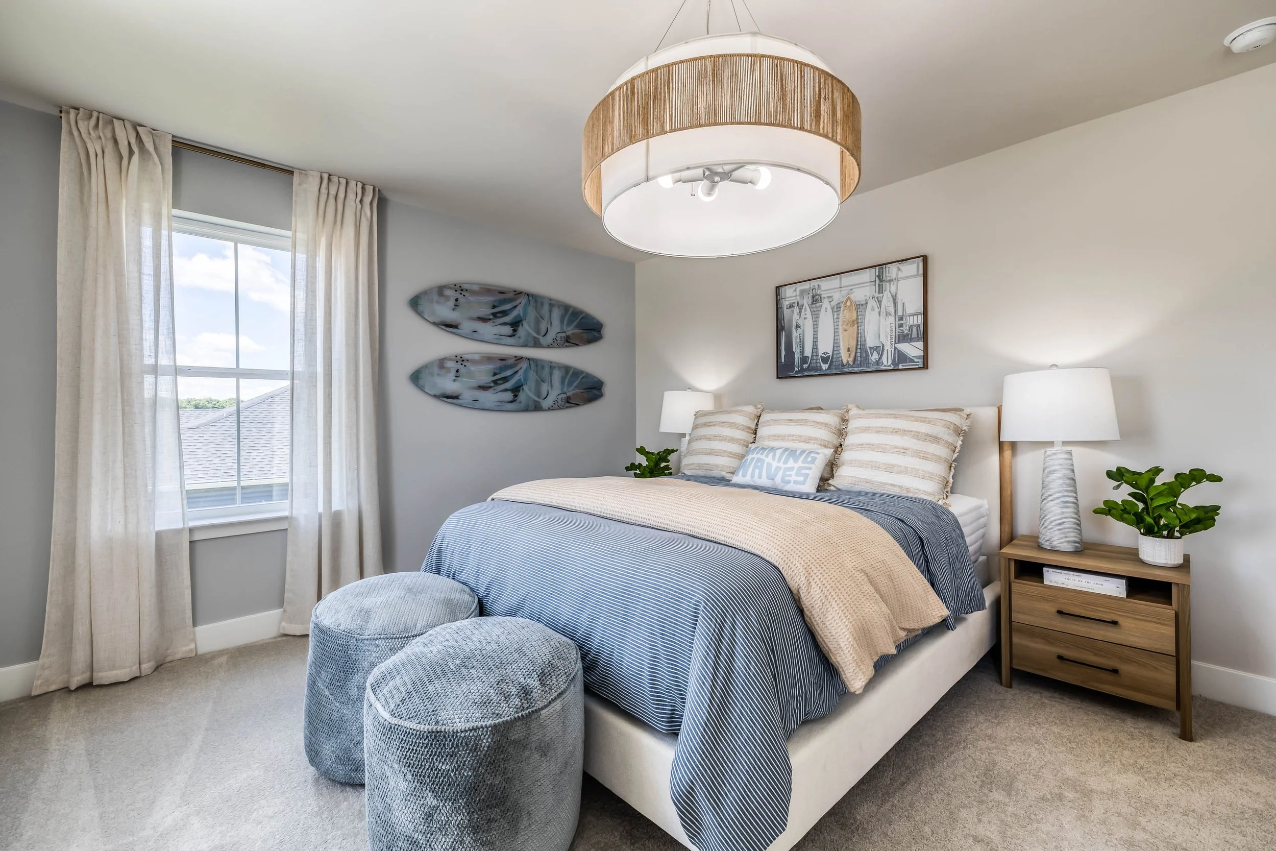 A cozy bedroom with a large bed featuring striped bedding and decorative pillows, two bedside lamps, a wooden nightstand, a window with sheer curtains, wall art with surfboards, and a ceiling light fixture with a fabric shade.