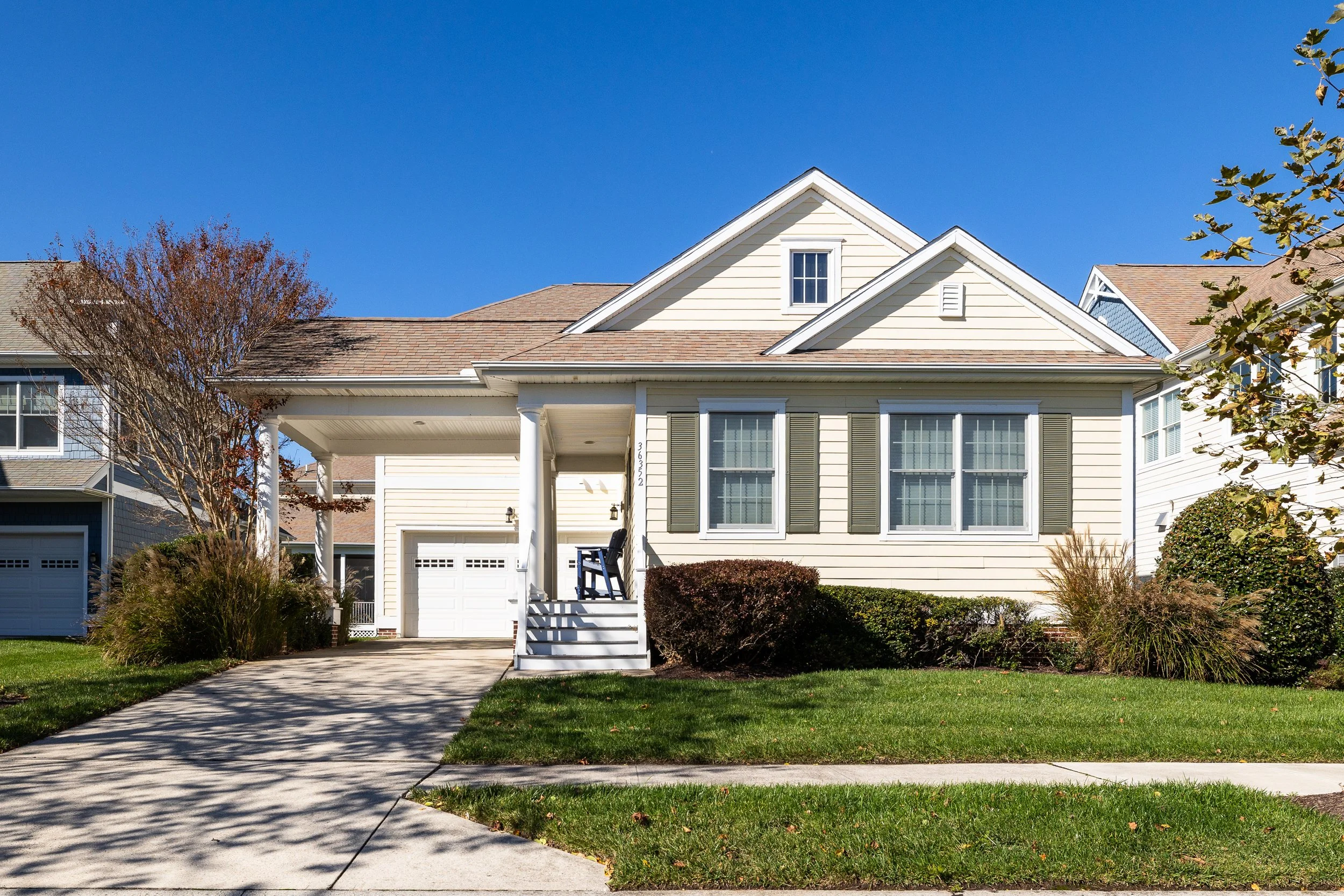 A two-story house with white siding, shutters, and a front porch, situated on a neatly landscaped lawn with shrubs and trees, under a clear blue sky.