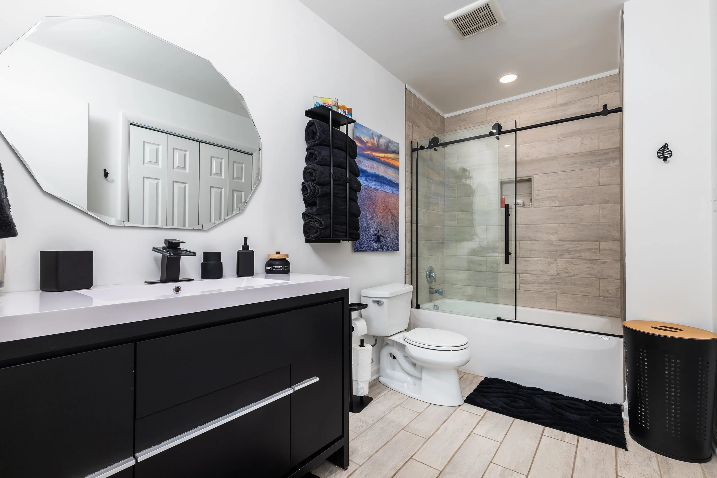 Modern bathroom with black vanity, white countertop, and black accessories, a toilet, a glass-enclosed shower with wood-look tiles, wall-mounted towel shelf with black towels, a large landscape painting, a black laundry basket, and a black rug.