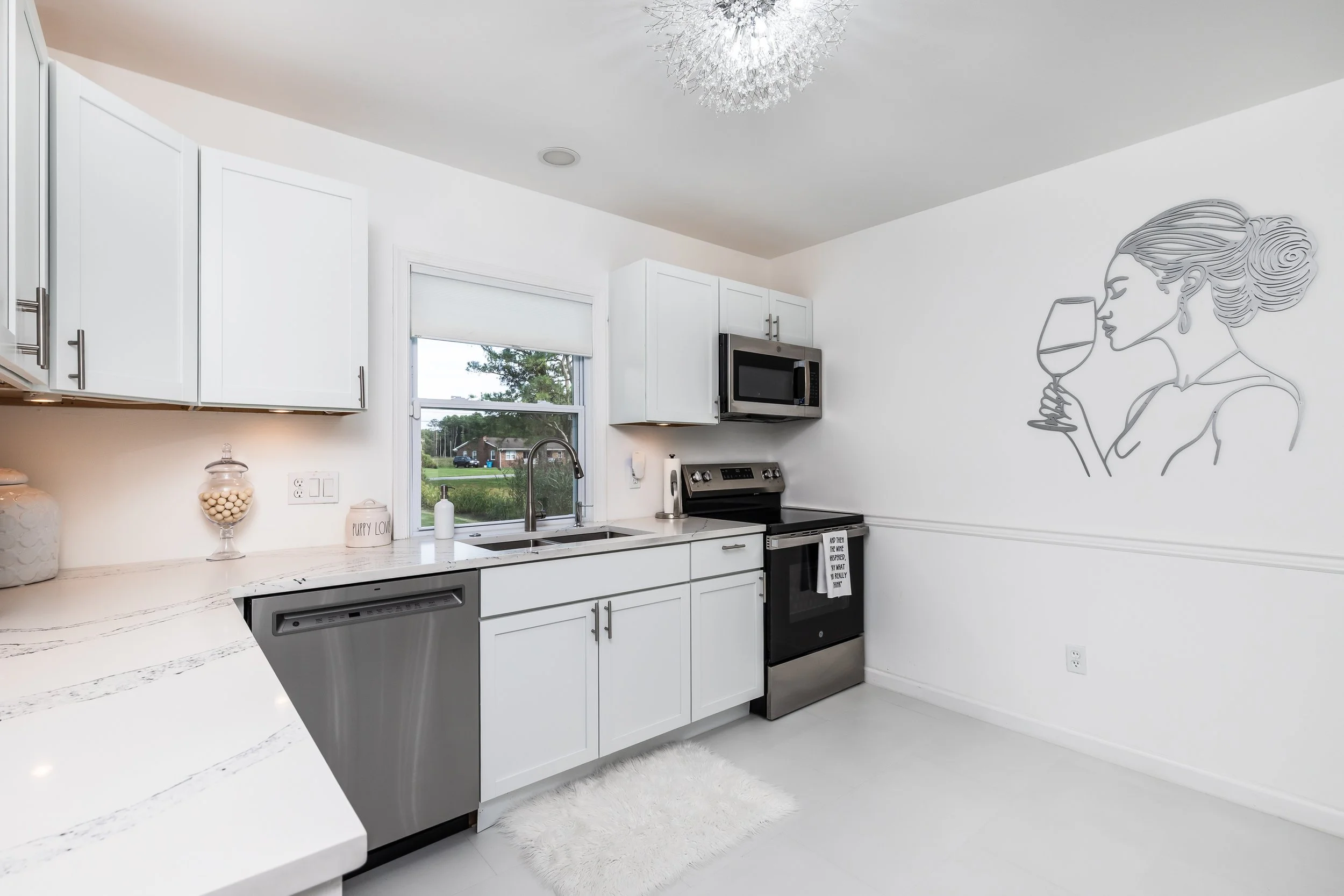 Kitchen with white cabinets, a window, gray and black appliances, a wall mural of a woman with her hair in a bun drinking wine, and a white fluffy rug.