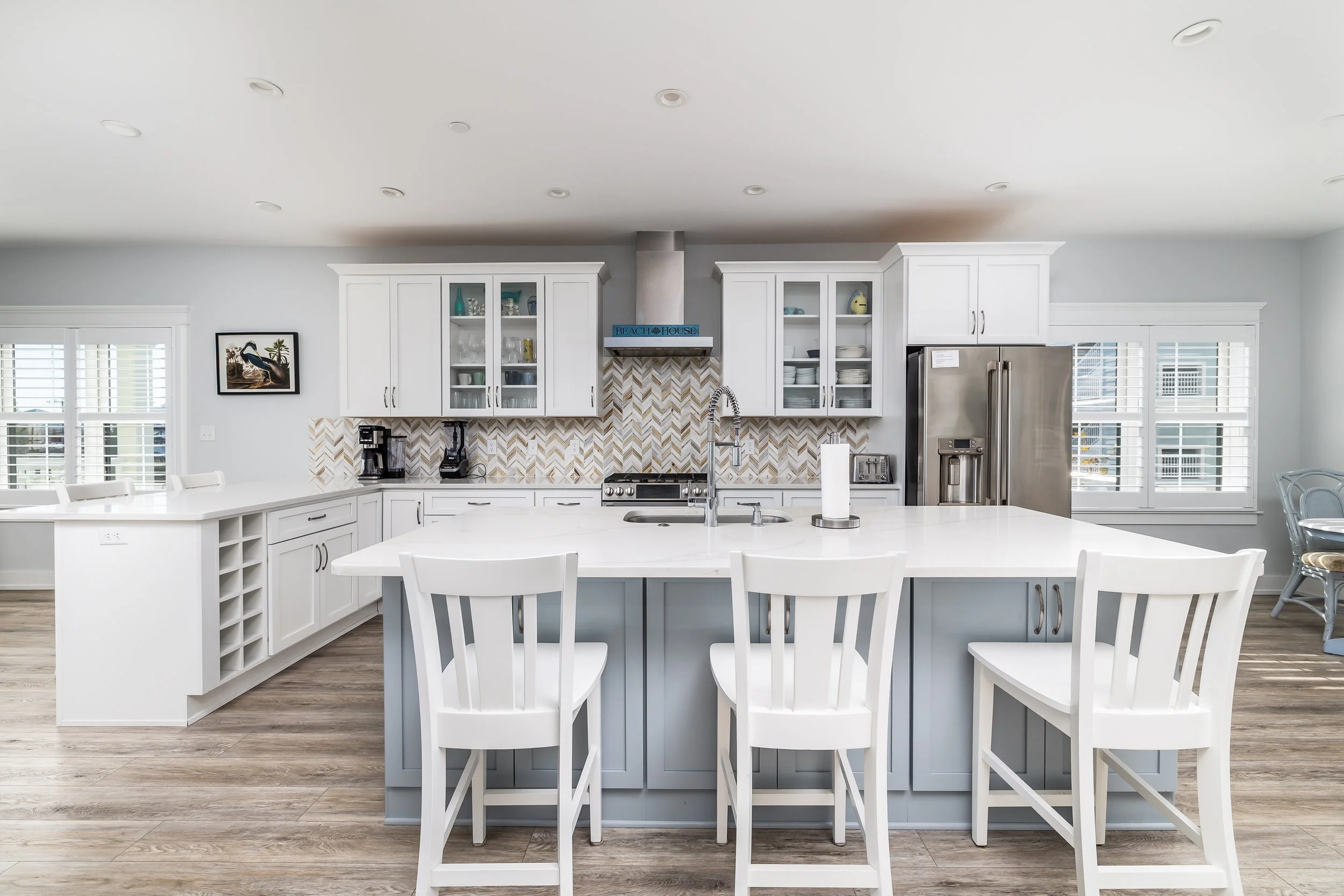 Modern kitchen with white cabinets, a large island with white chairs, stainless steel appliances, and a patterned tile backsplash.