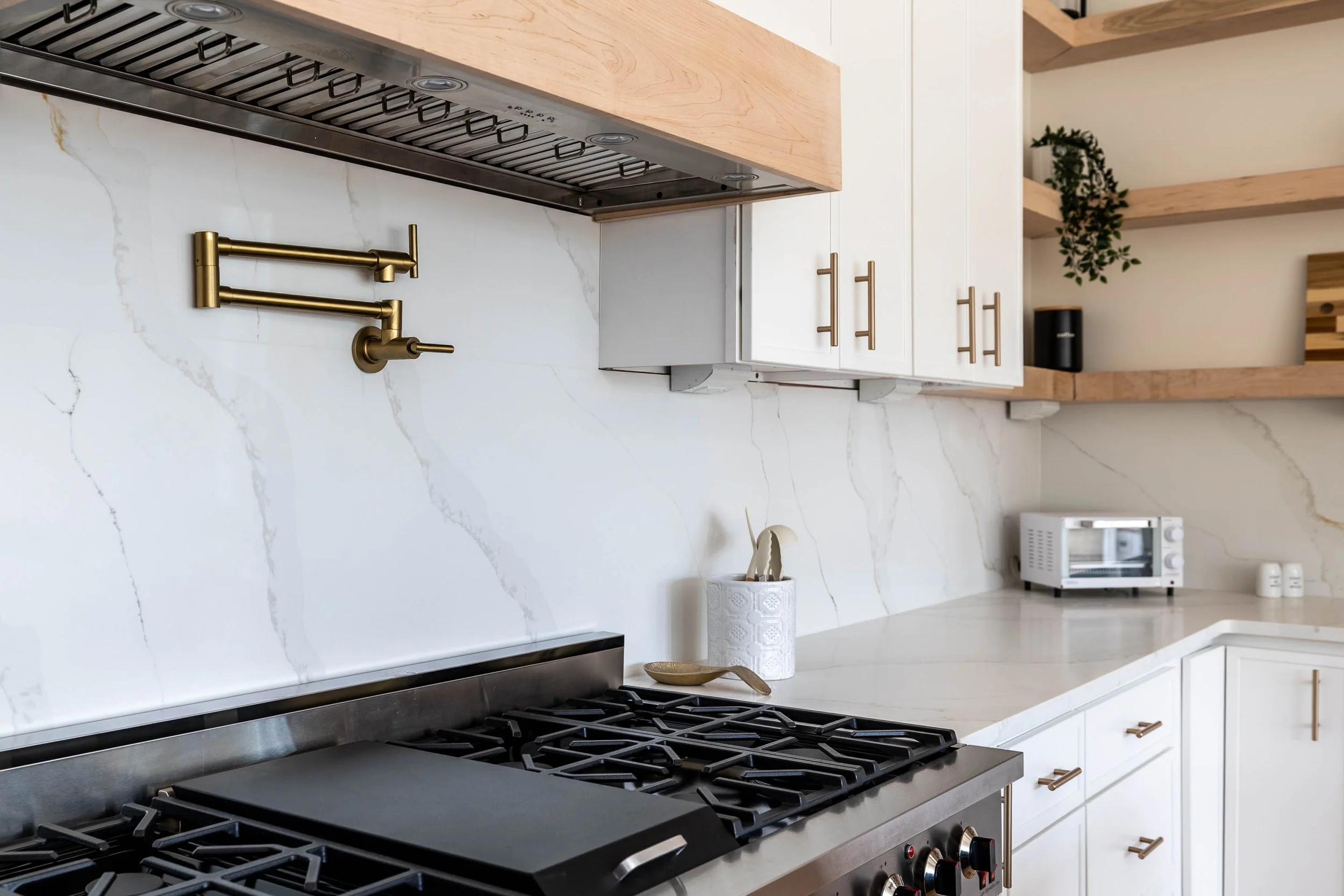 A modern kitchen with a gas stove, white cabinets with gold handles, a white marble backsplash, and wooden open shelves with decorative items and greenery.
