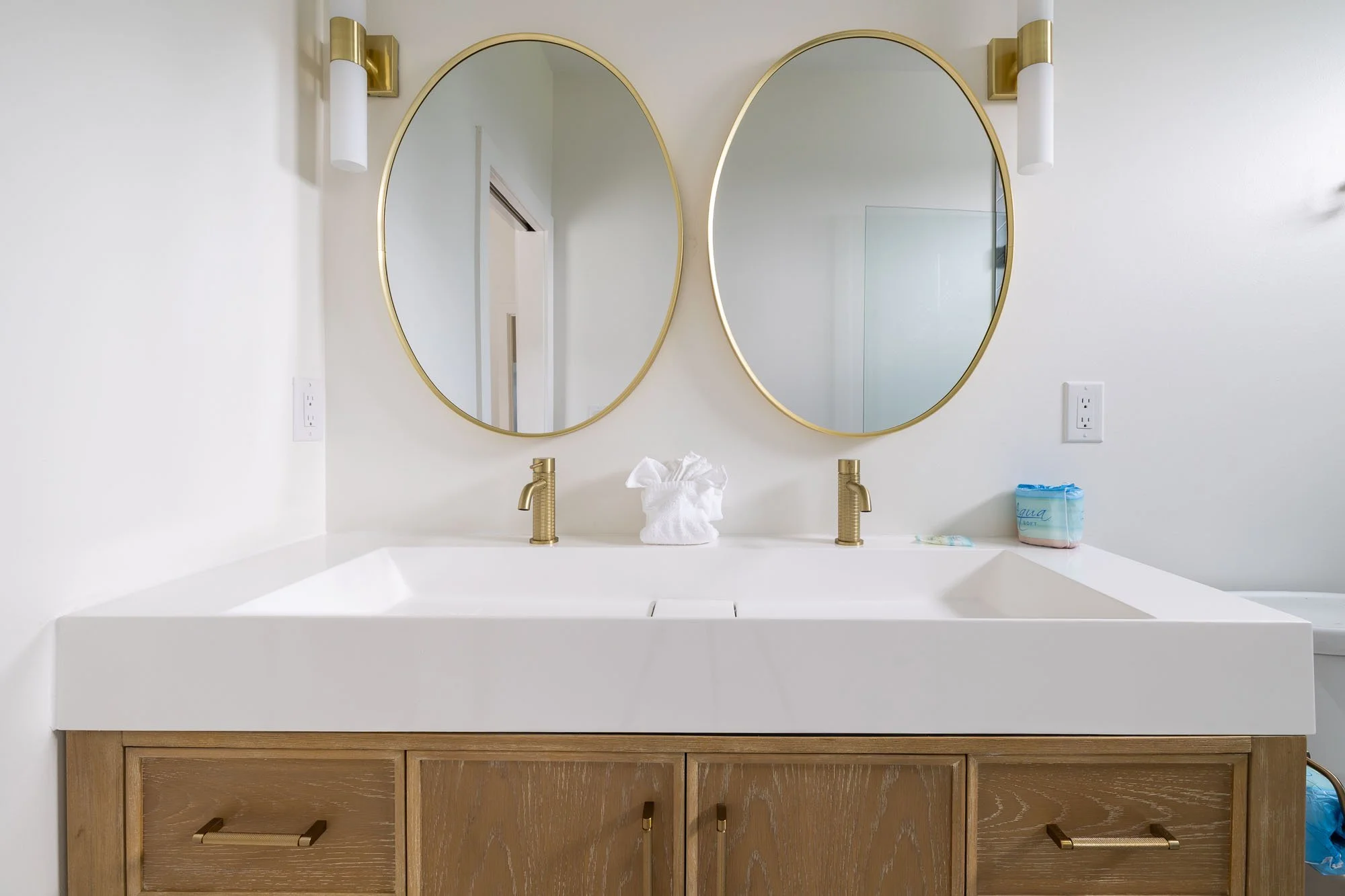 Bathroom vanity with two oval mirrors, gold fixtures, and a wooden cabinet under a white countertop with a towel and toiletries.
