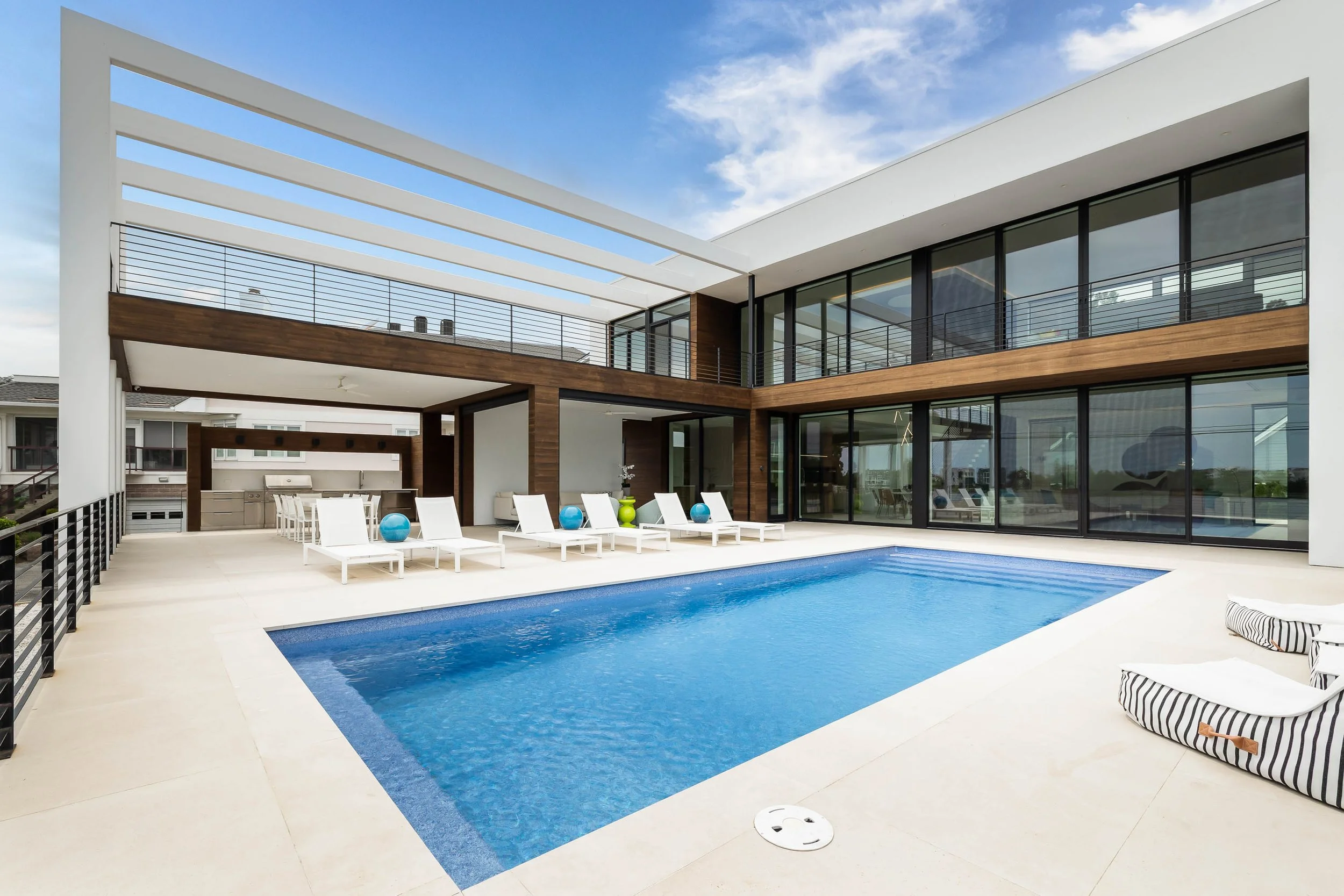 Modern house with large glass windows and a swimming pool, outdoor seating with white lounge chairs and blue and green decorative balls, balcony with black railing, partly cloudy sky.
