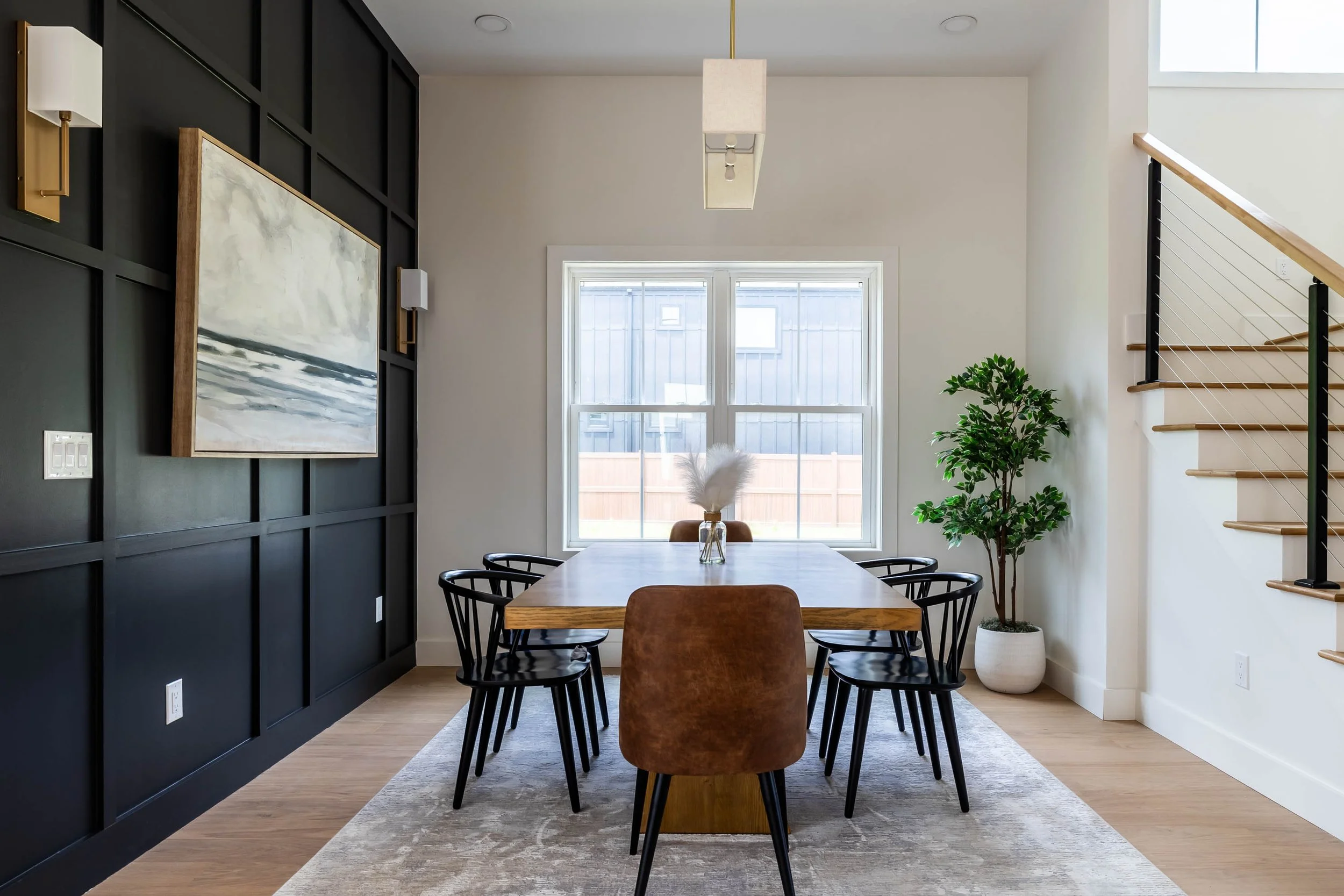Modern dining room with a wooden table, six black chairs, a large window, a potted plant, and a staircase with black railing.