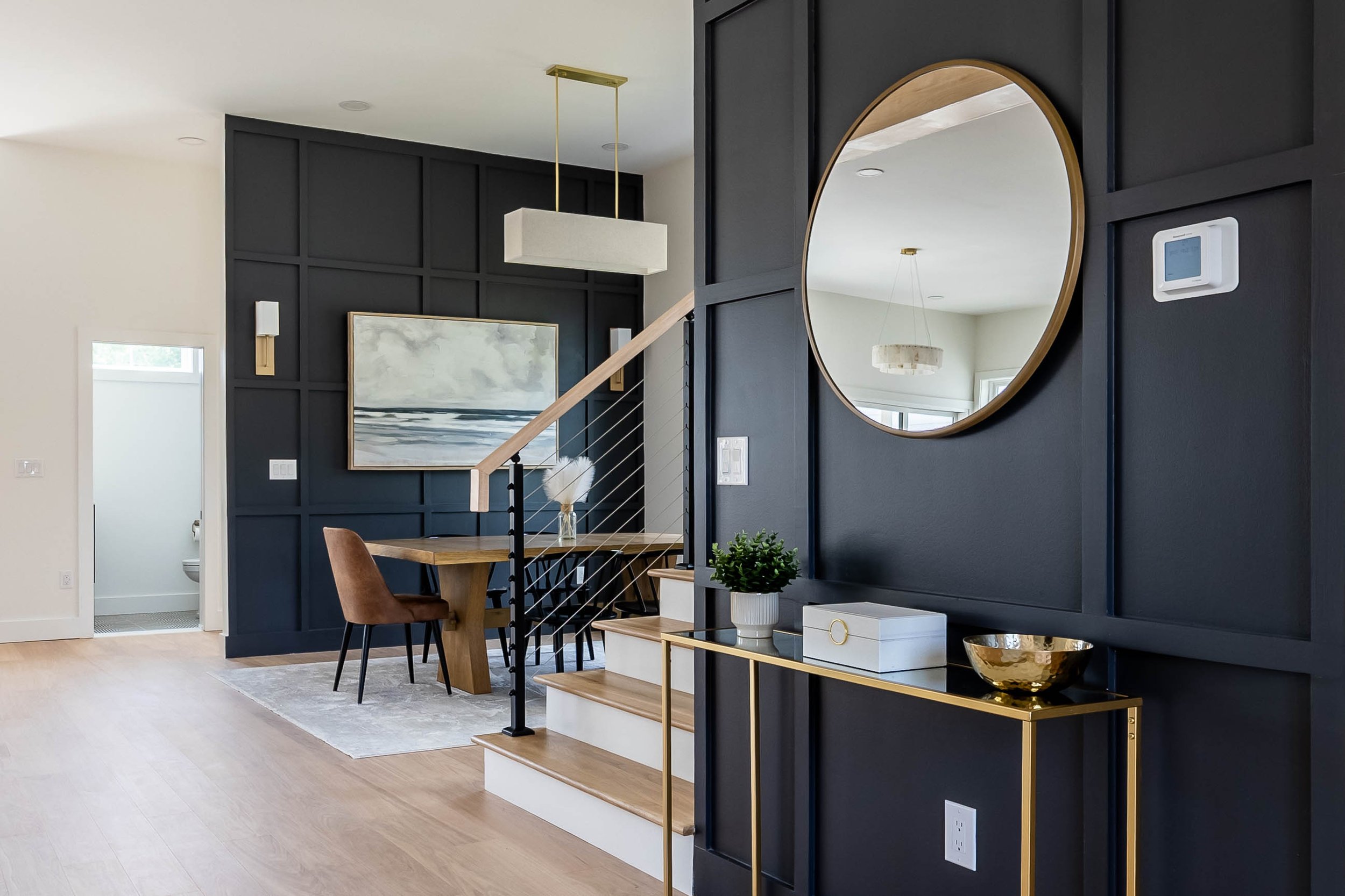 Interior view of a modern living and dining area with black wall paneling, a wooden table, brown chairs, a large digital thermostat on the wall, a round mirror, and a gold accent console table with decorative bowls and a plant.