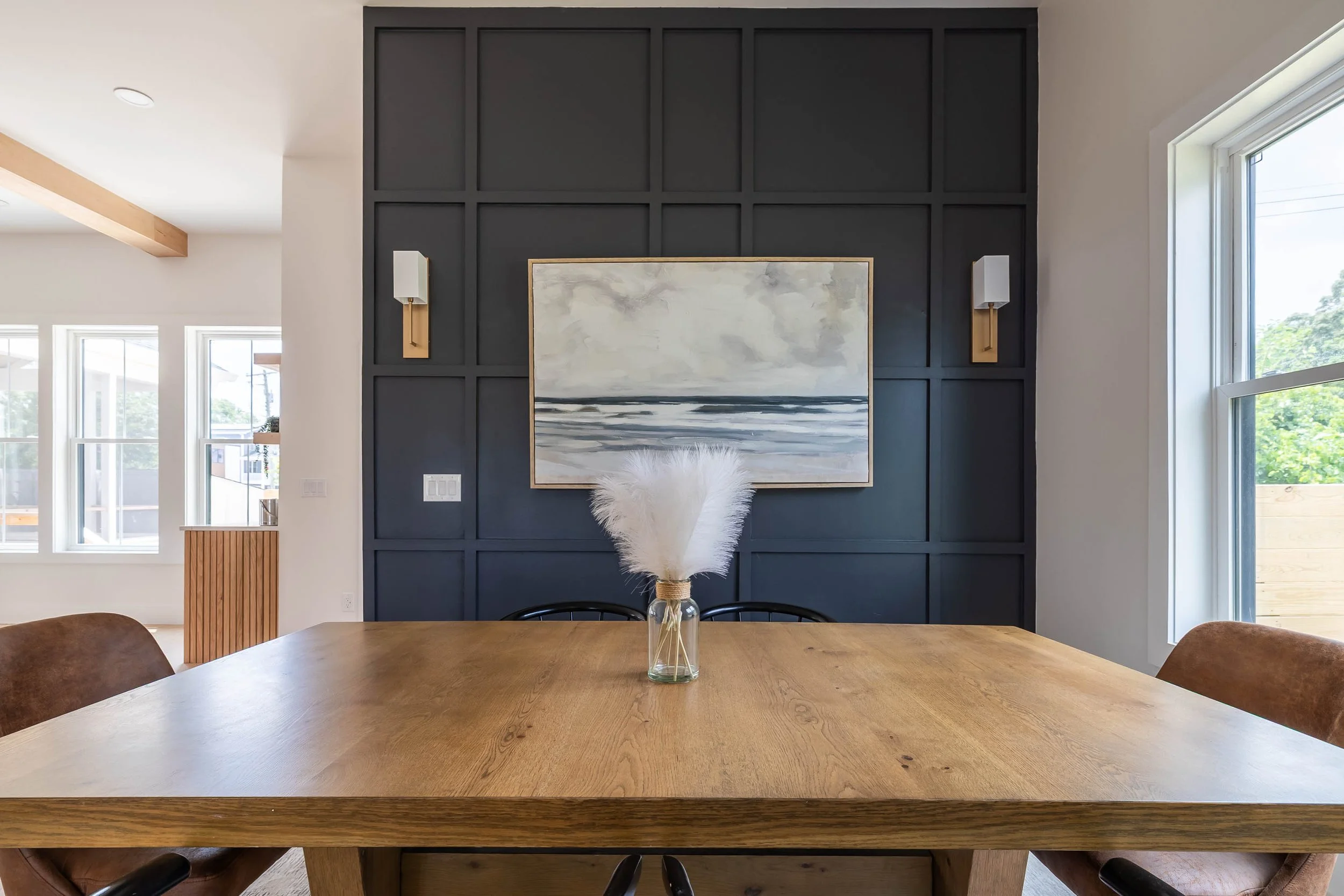 View of dining room with wooden table, a vase with white pampas grass, black chairs, black paneled wall with abstract painting, wall sconces, large windows, and natural light.