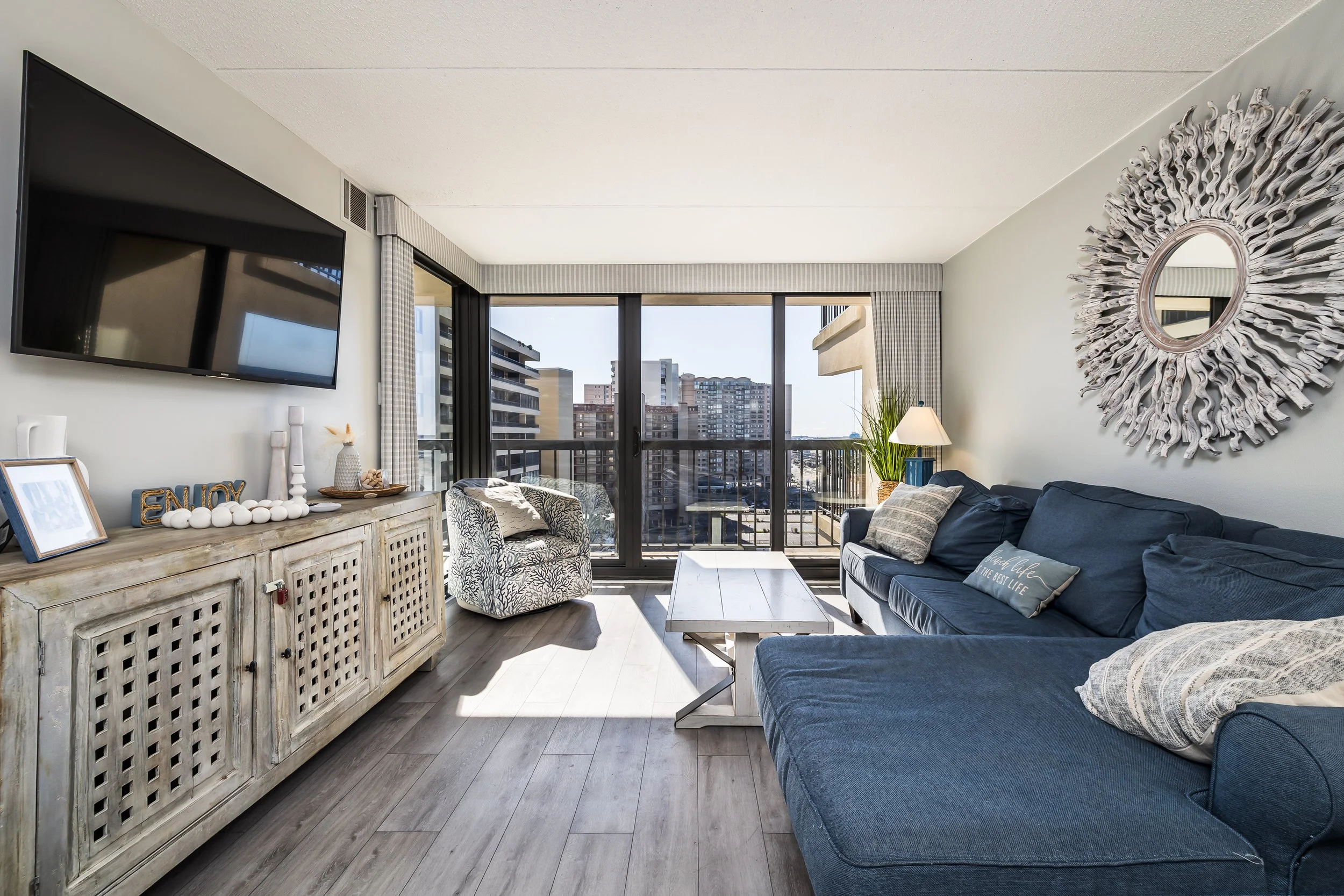 Living room with large window and balcony, blue sectional sofa with throw pillows, patterned armchair, wooden coffee table, wall-mounted TV, decorated sideboard, decorative mirror on wall, potted plant, lamp, bright daylight.