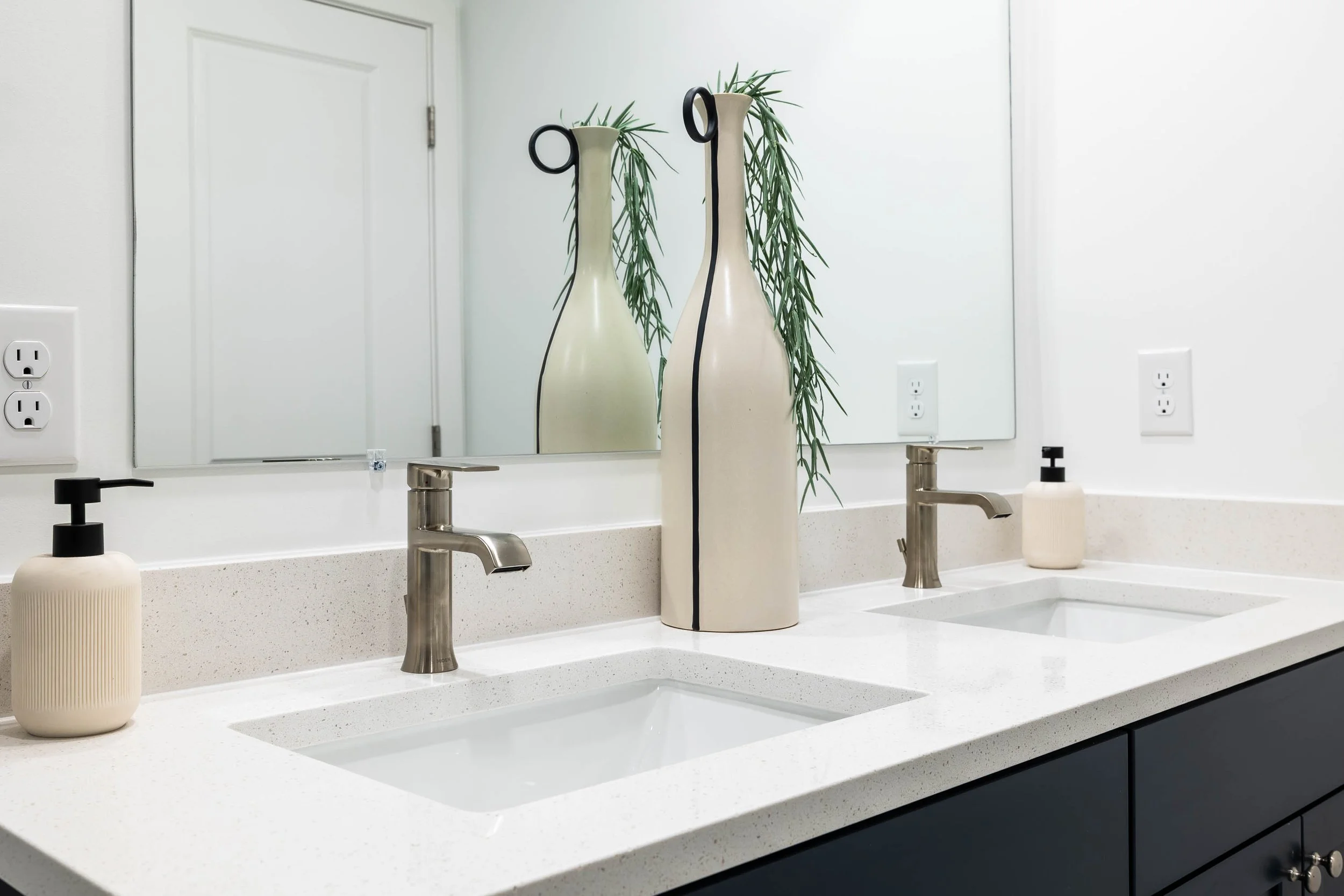 Modern bathroom vanity with two faucets, a large mirror, a soap dispenser, and decorative vases with greenery on a speckled white countertop.