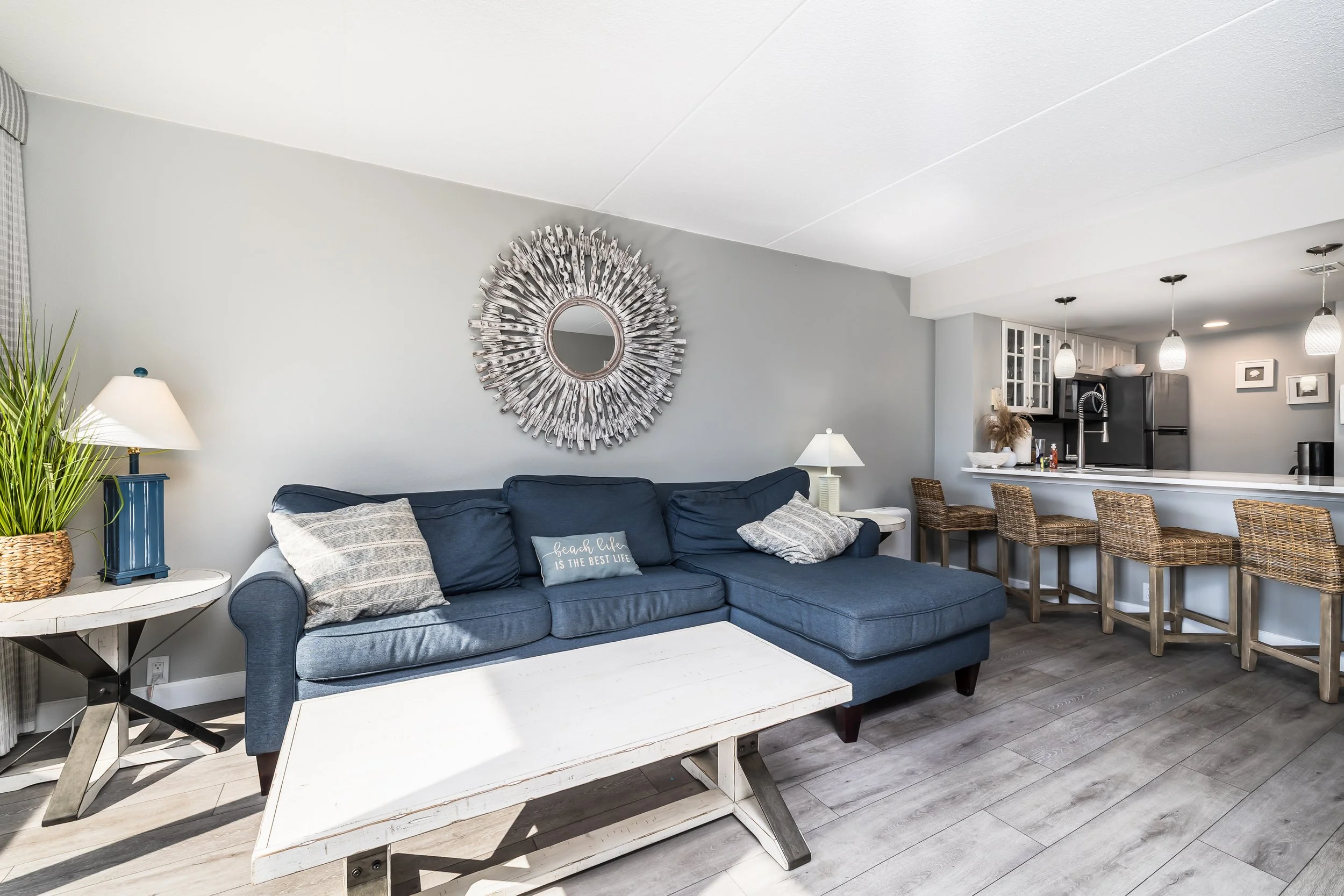 Living room with a blue sectional sofa, a white coffee table, wicker and wood bar stools at kitchen counter, and modern decor including a sunburst mirror and potted plant.