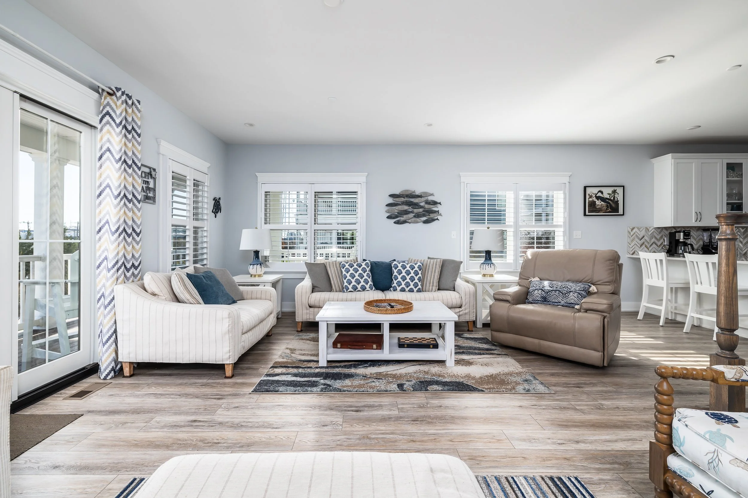 Living room with light blue walls, white trim, multiple windows with blinds, a sliding glass door with patterned curtains, and wooden flooring. Contains sofas, chairs, a white coffee table, lamps, framed artwork, and a nearby kitchen area with white 