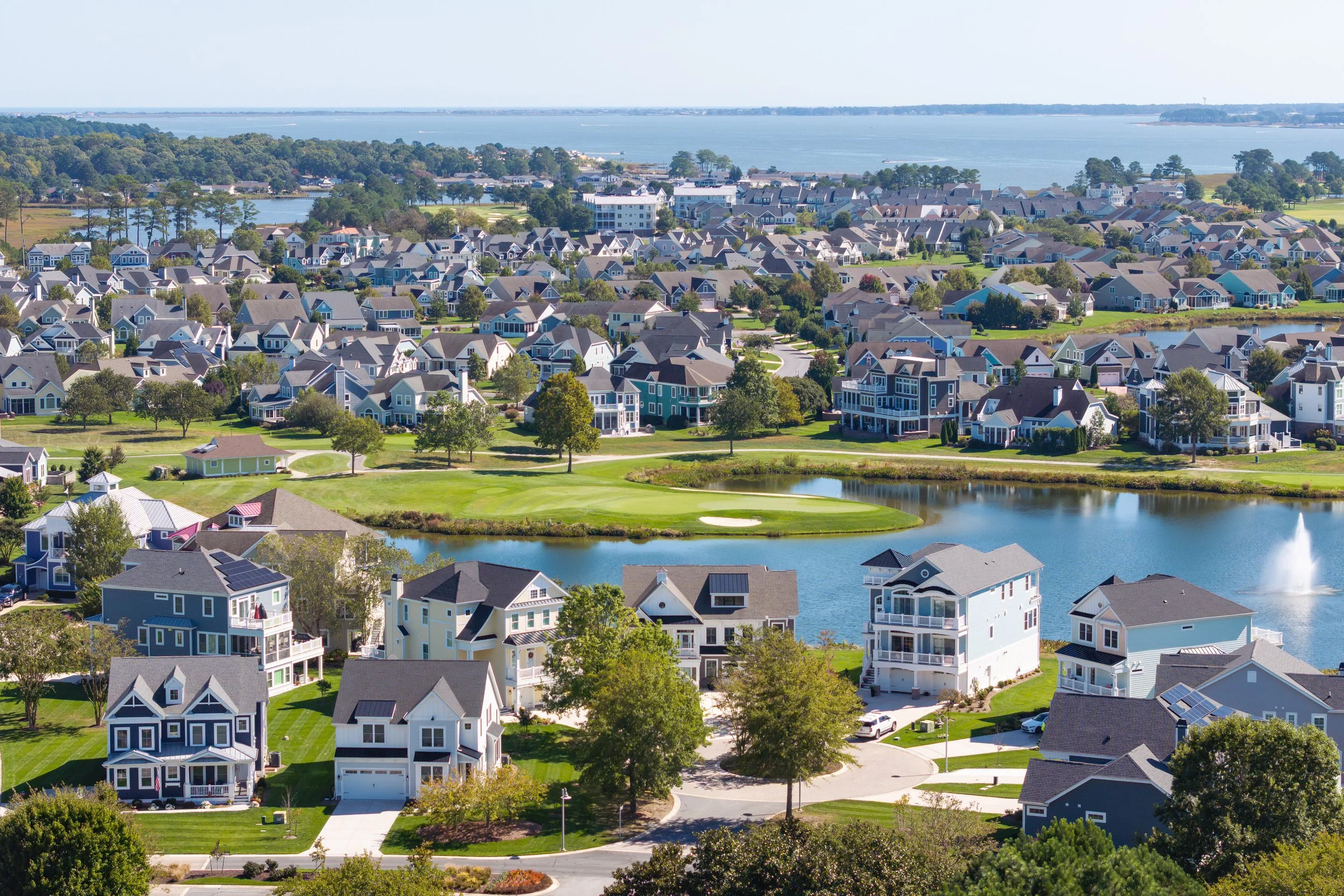 Aerial view of a neighborhood with houses around a pond, featuring a fountain in the water and a golf course at the back, near a large body of water.