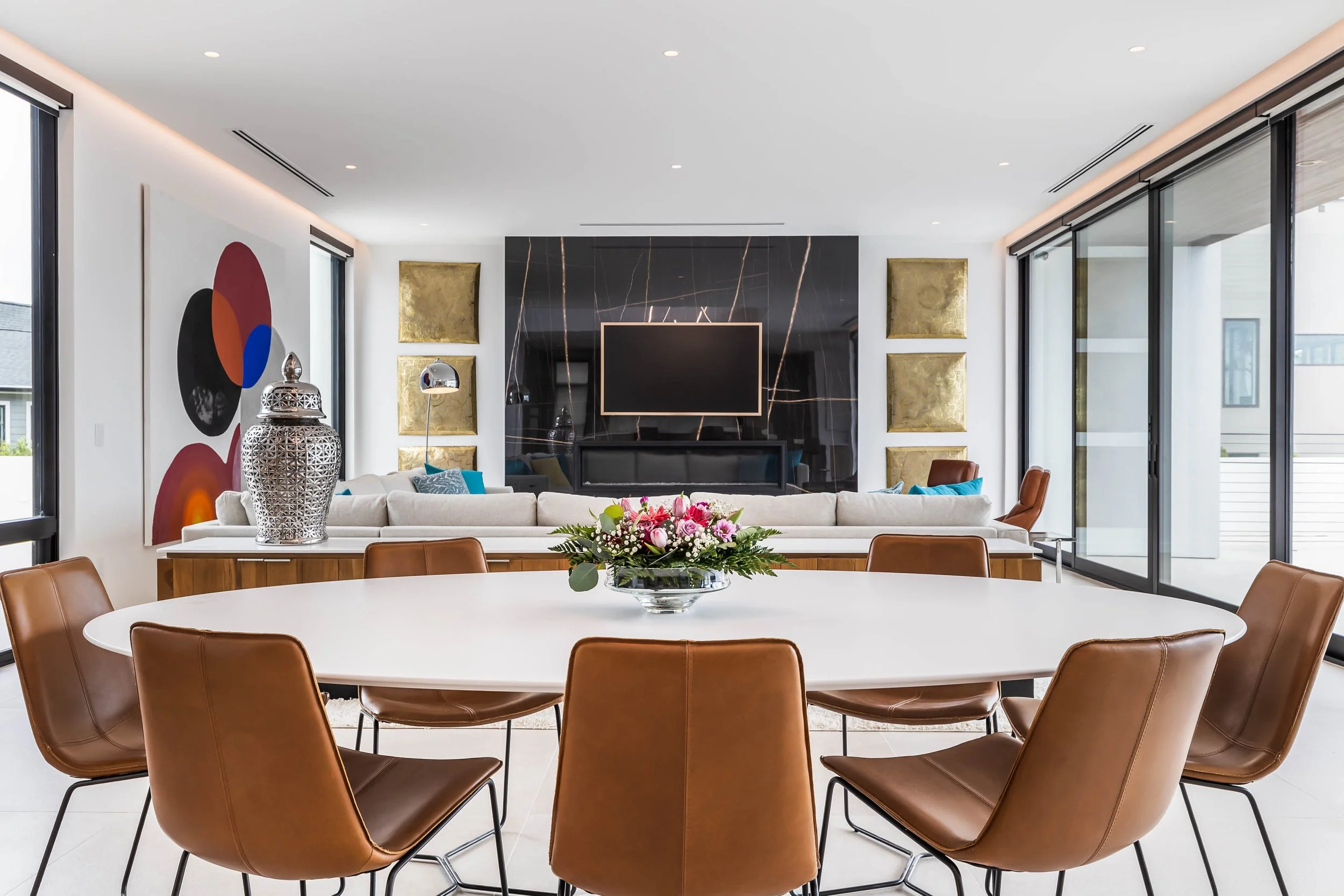 Modern living room with a black marble fireplace, wall art, a large TV, and a round dining table with leather chairs, a floral centerpiece, large windows, and sliding glass doors.