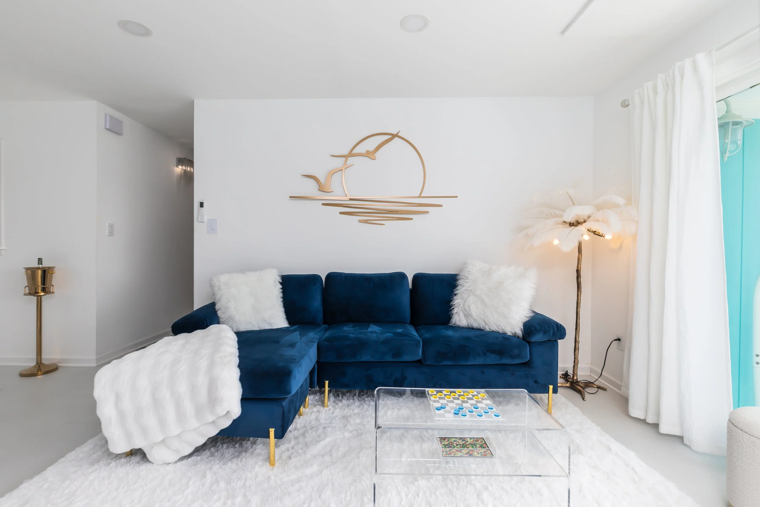 Living room with a navy blue sofa, white fluffy pillows, a fluffy blanket, a white shag rug, a clear acrylic coffee table with a board game, a tall feather lamp, white curtains, and a minimalist wall art of a sunset with birds in gold.