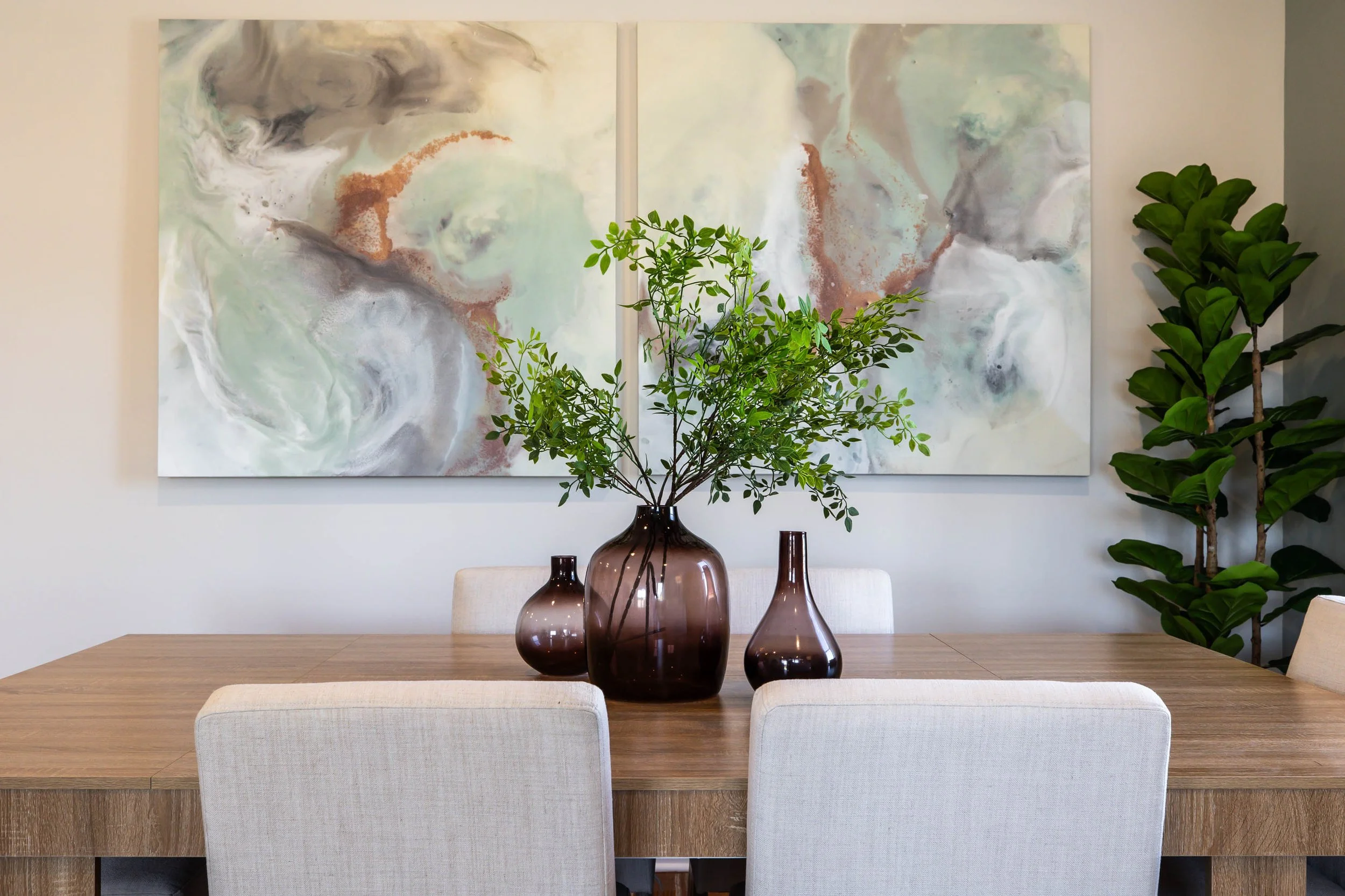 A modern dining room features a wooden table with three brown glass vases and a potted green plant. Behind the table, a pair of abstract watercolor paintings with pastel colors are mounted on the wall, flanked by two green plants on each side.