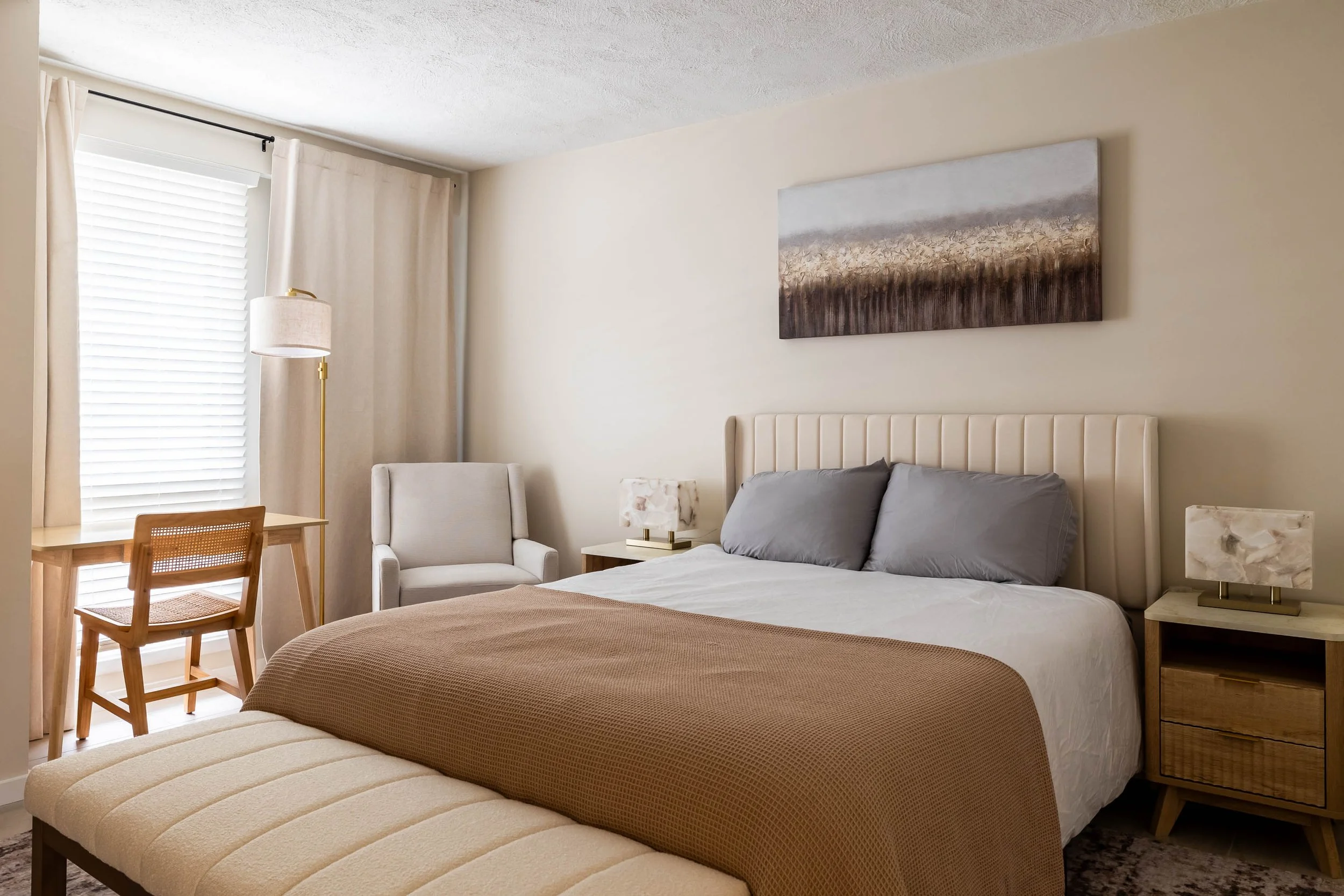 A bedroom with a bed, nightstand, chairs, and wall art, illuminated by natural light from a window with blinds and curtains.