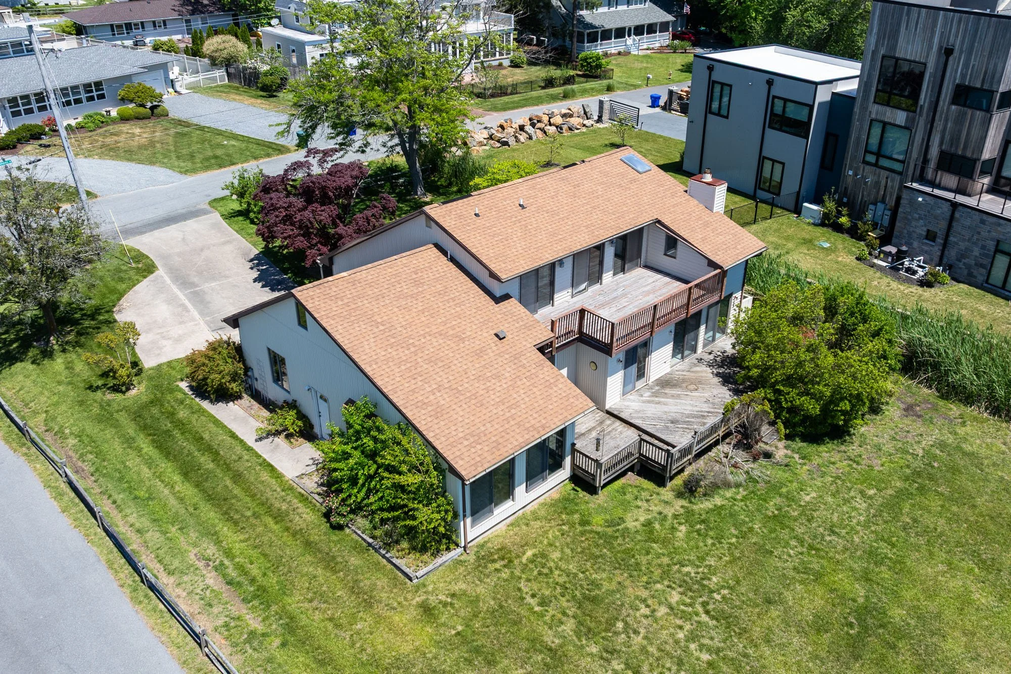 Aerial view of a residential neighborhood featuring a mix of houses with different styles, including a house with a brown-tiled roof, a modern multi-story house with black windows and wooden siding, and smaller houses with white siding. The yards are landscaped with trees, bushes, and grass, and there are driveways, sidewalks, and cars visible. The area is lush with greenery and appears to be in spring or early summer.