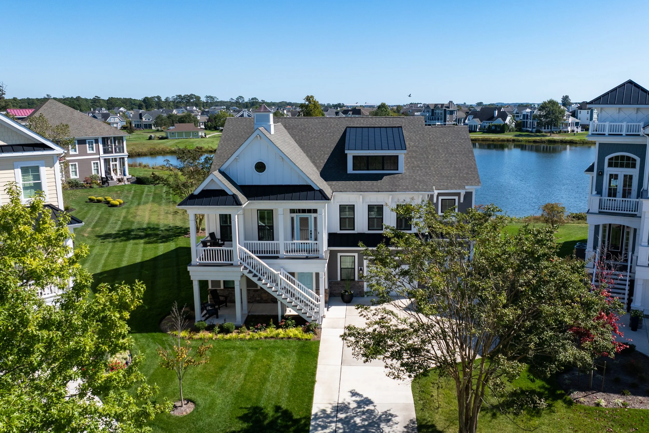 A white and gray two-story house with a porch and stairs, surrounded by green lawns, trees, and neighboring houses near a lake.