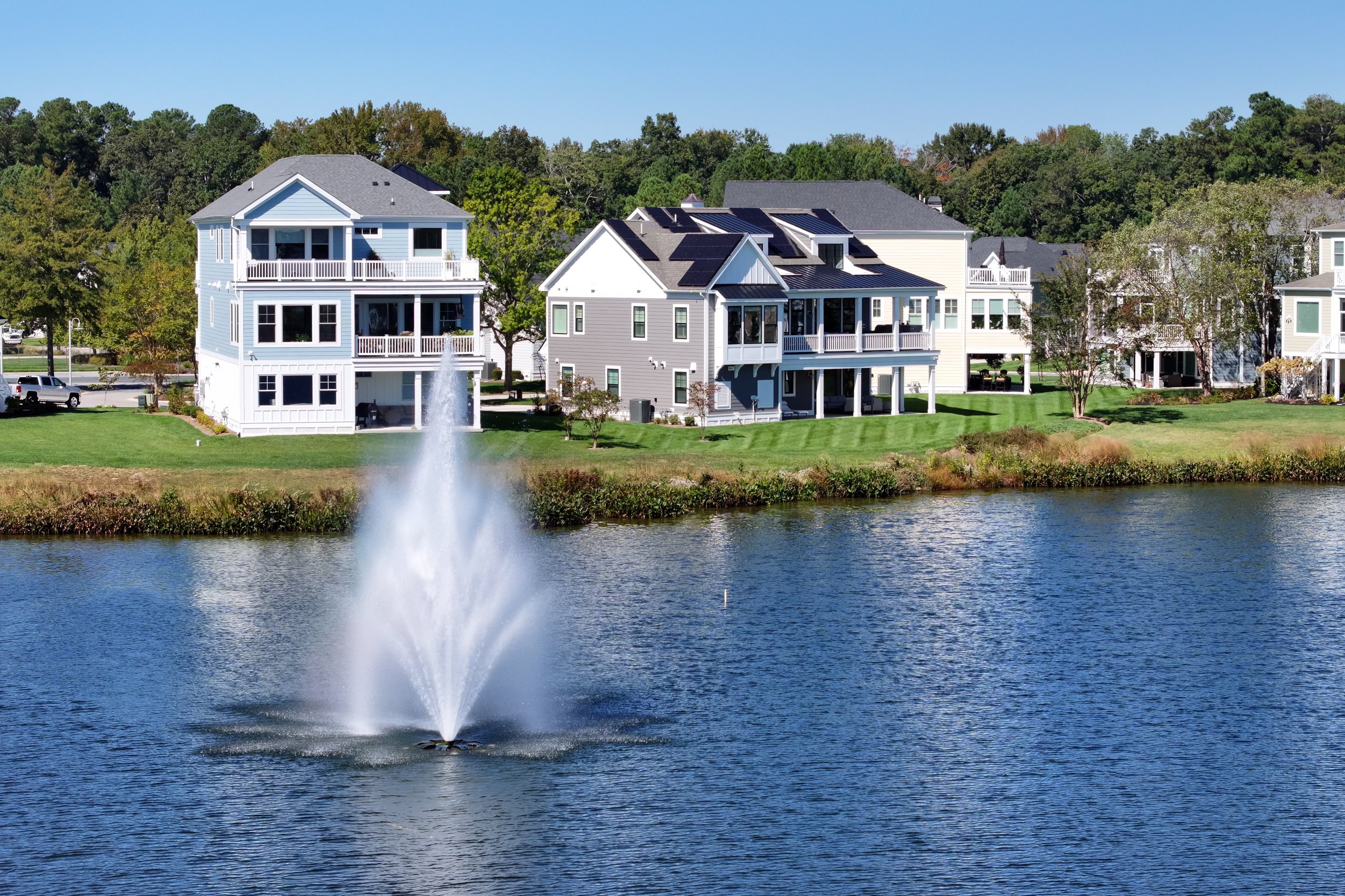 A lakeside residential area featuring three multi-story houses with lawns and trees, with a large fountain spraying water in the lake in the foreground, and a clear blue sky overhead.