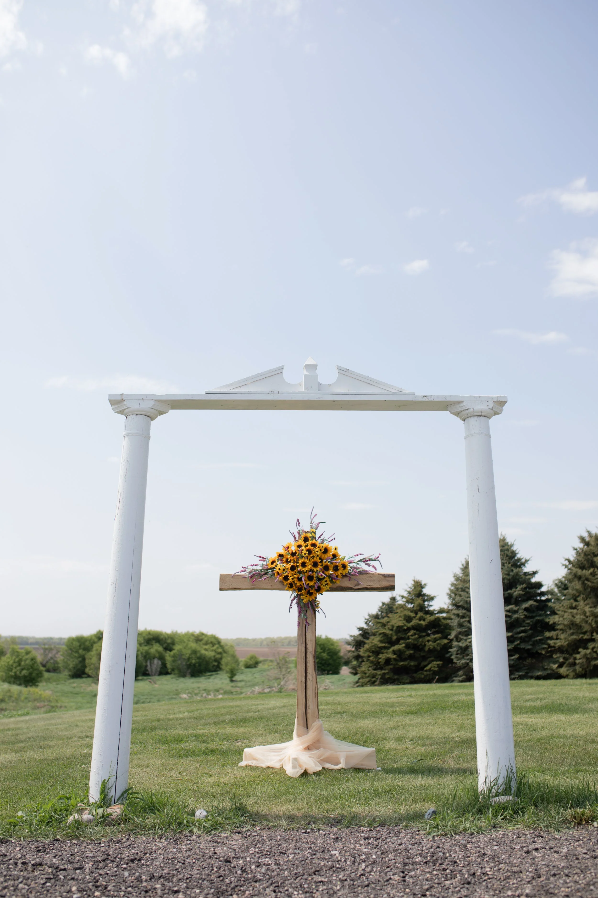 A white wooden arch with columns and a triangular pediment, decorated with a bouquet of sunflowers and purple flowers, set outdoors in a grassy area, likely for a wedding ceremony.