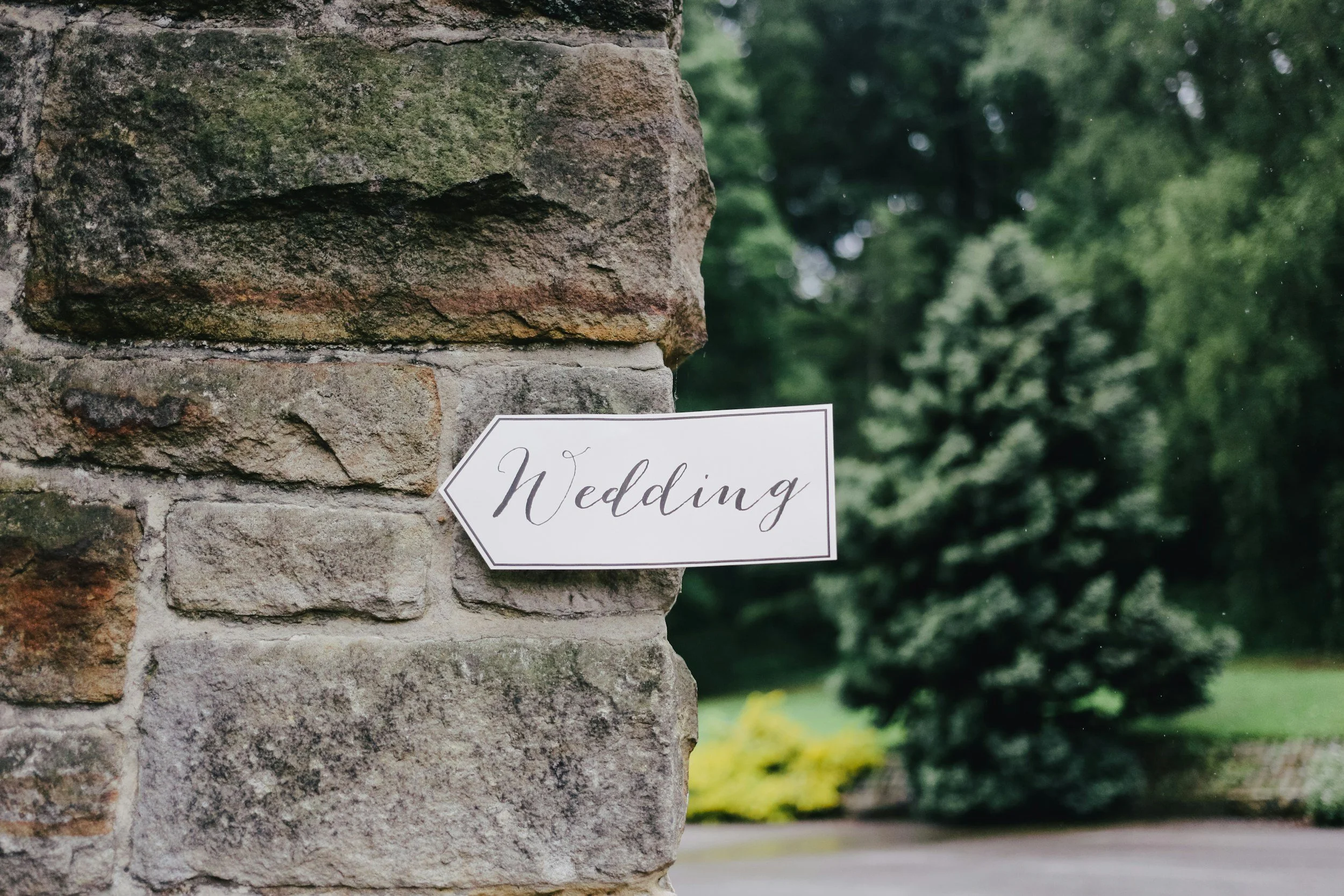 Close-up of a stone wall with a sign that partially reads 'Wedding' in elegant script, with blurred trees and greenery in the background.