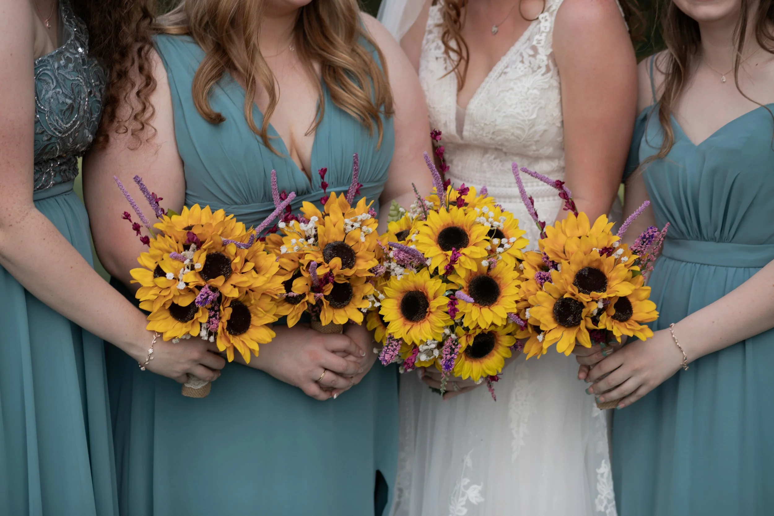 Close-up of four women wearing teal and white dresses, holding a large bouquet of sunflowers and purple, white, and pink flowers, at a formal event.