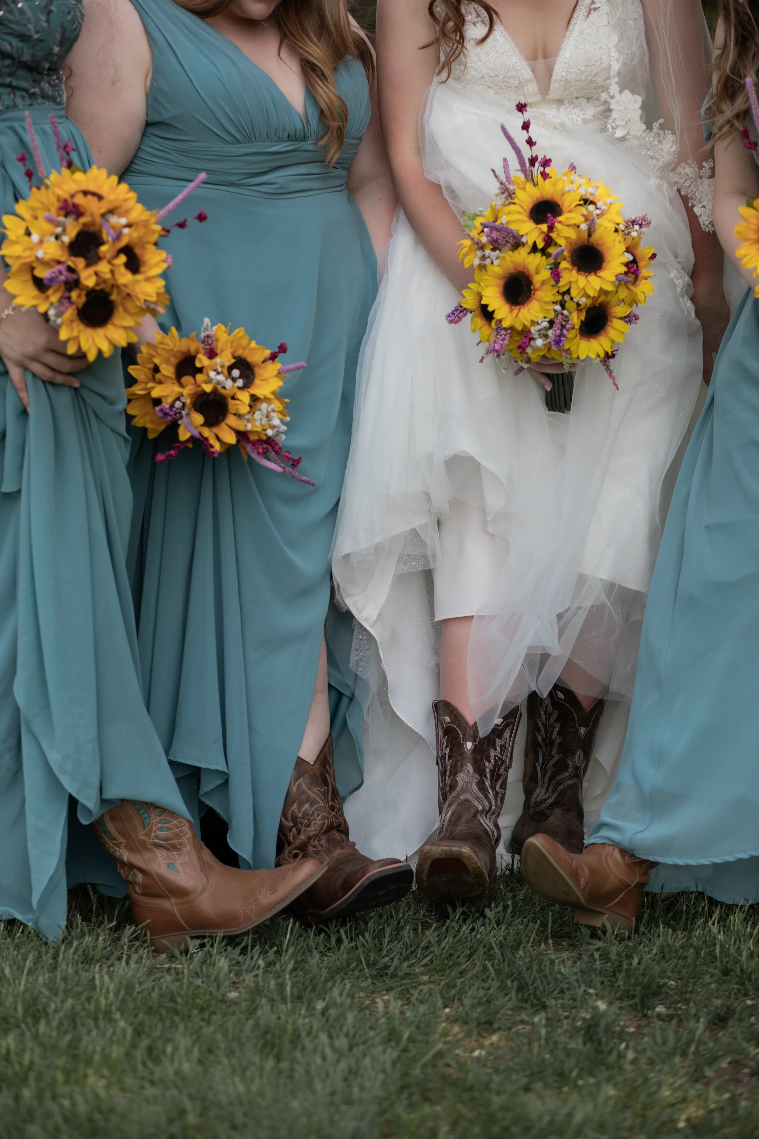 Close-up of women wearing cowboy boots, holding sunflowers, at a wedding or celebration, with grass on the ground.