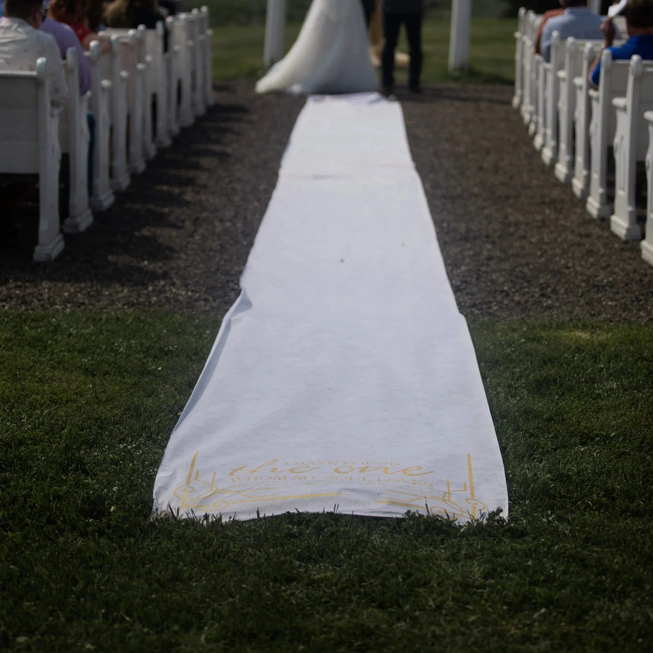 A white cloth table runner with decorative gold lettering and designs, laid on grass, at an outdoor event with white chairs arranged in rows on both sides.