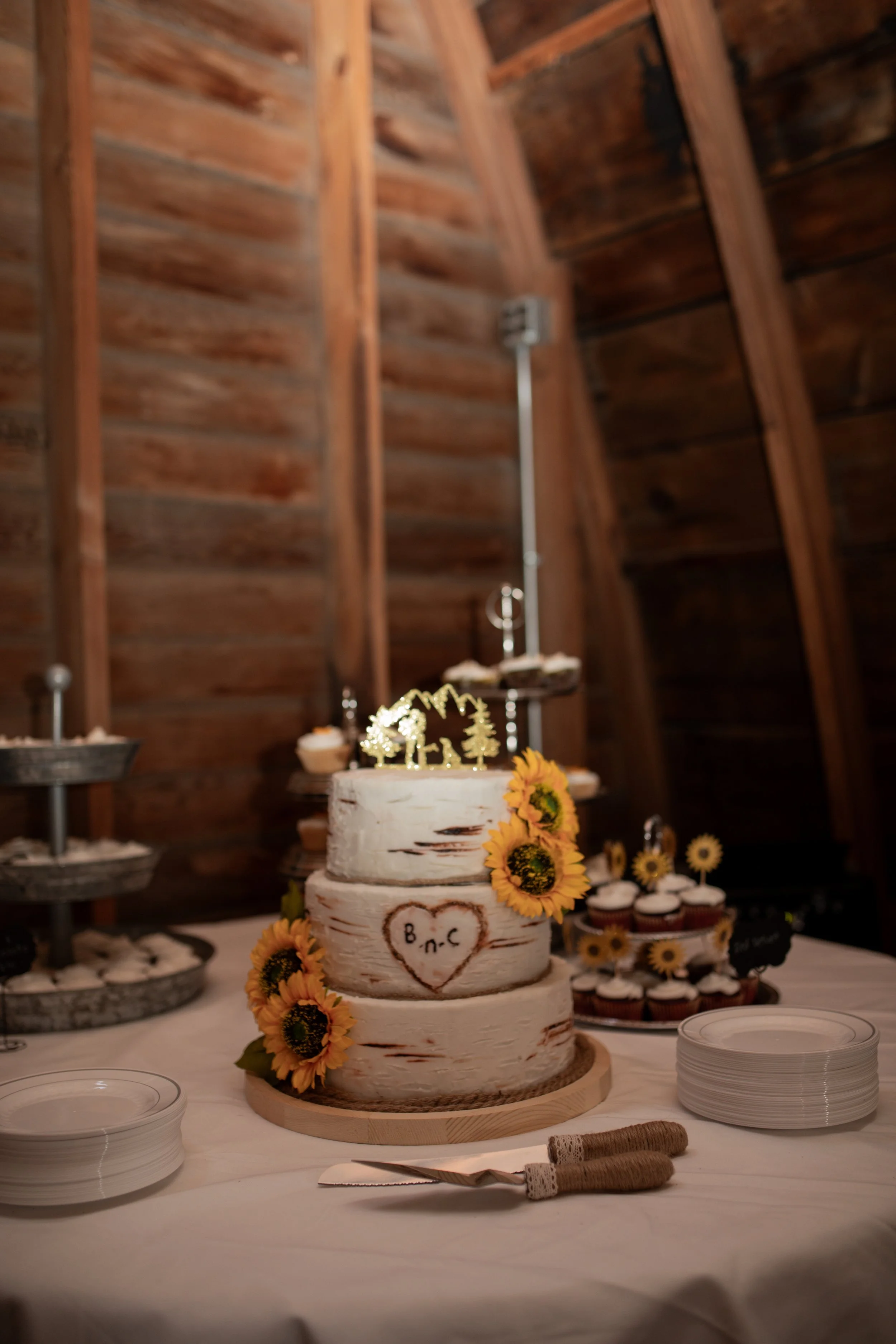 A rustic wooden cake with sunflower decorations and a small gold topper, sitting on a table with white plates and utensils, at a celebration or wedding event.
