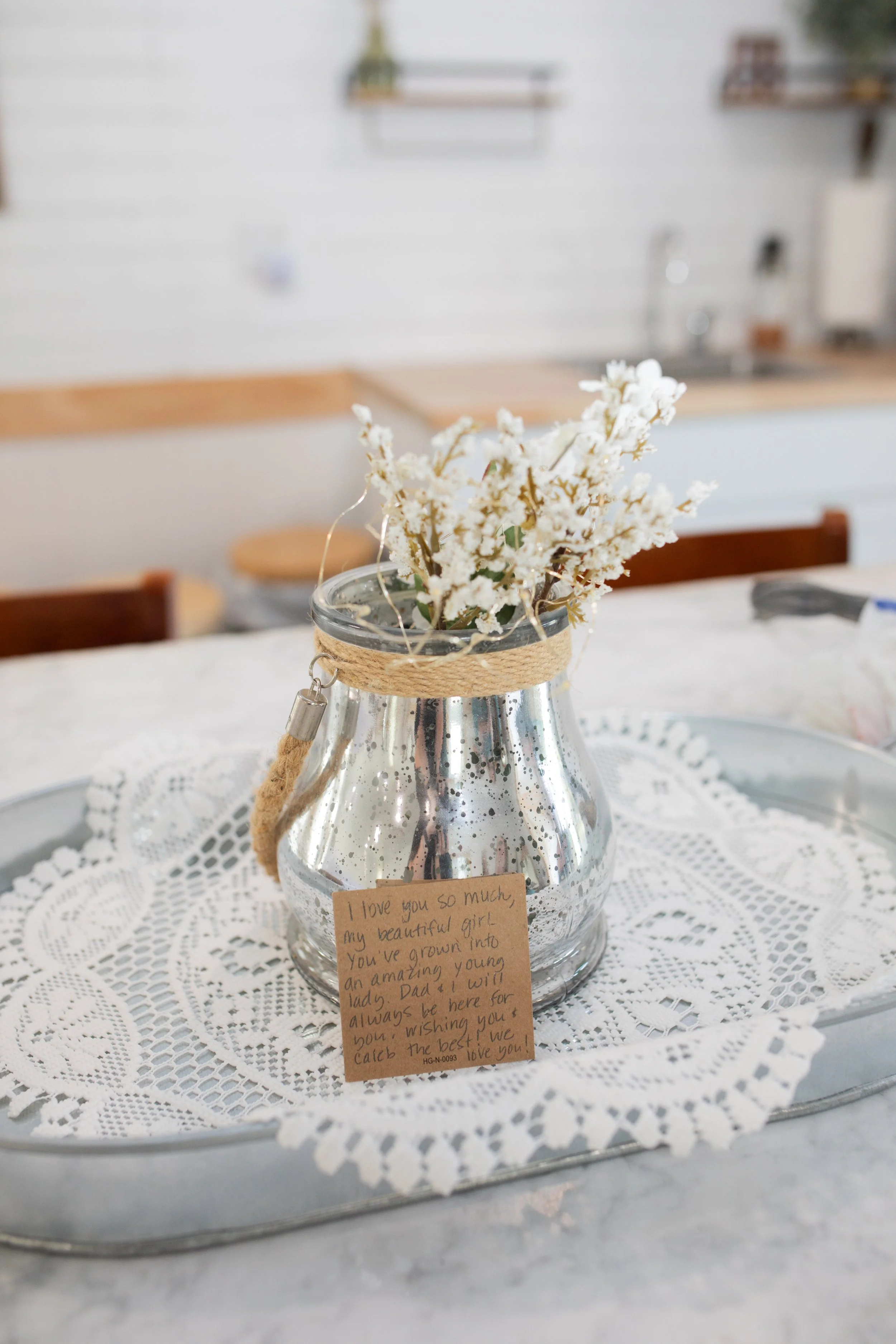A small glass vase with white artificial flowers, decorated with a jute string, placed on a white lace tablecloth. There is a handwritten note attached to the vase expressing love and pride for a girl who has grown into an amazing young lady.