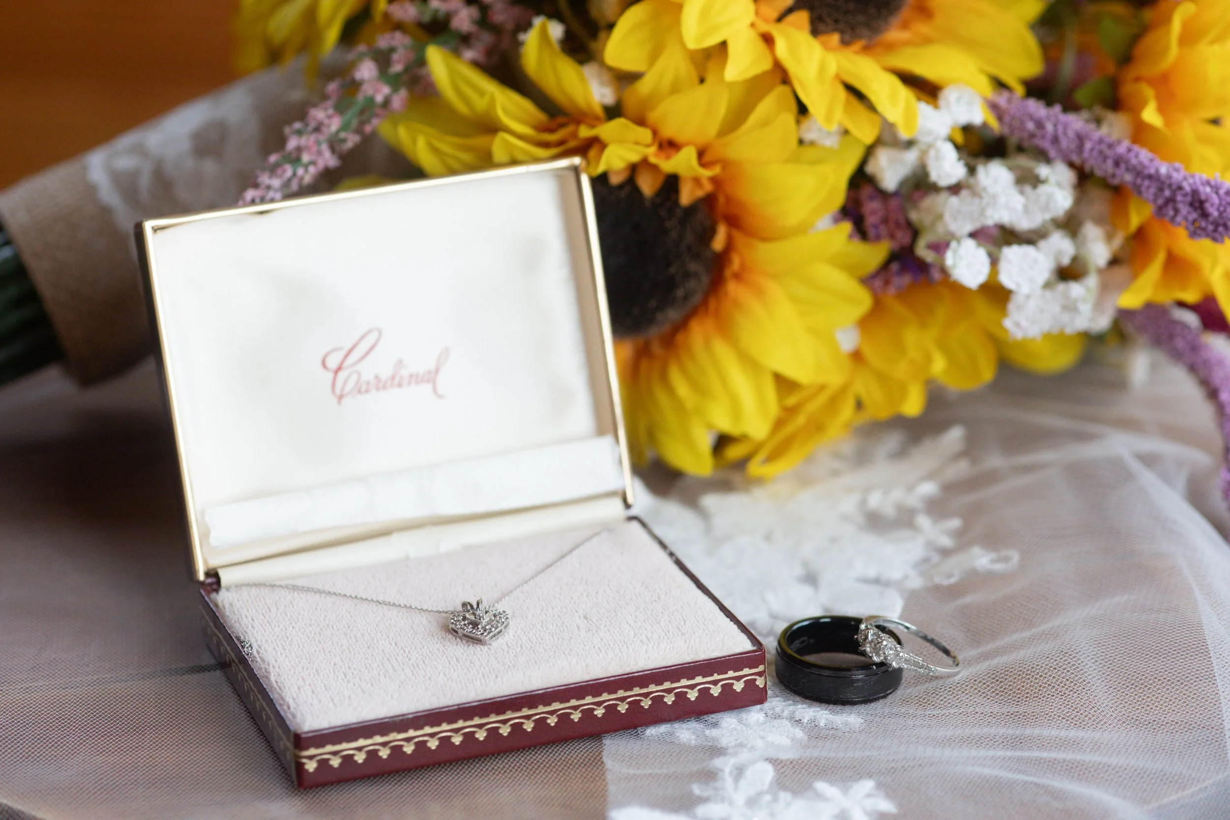 Open jewelry box with a silver ring inside, placed on a table with a bouquet of yellow, purple, and white flowers in the background.