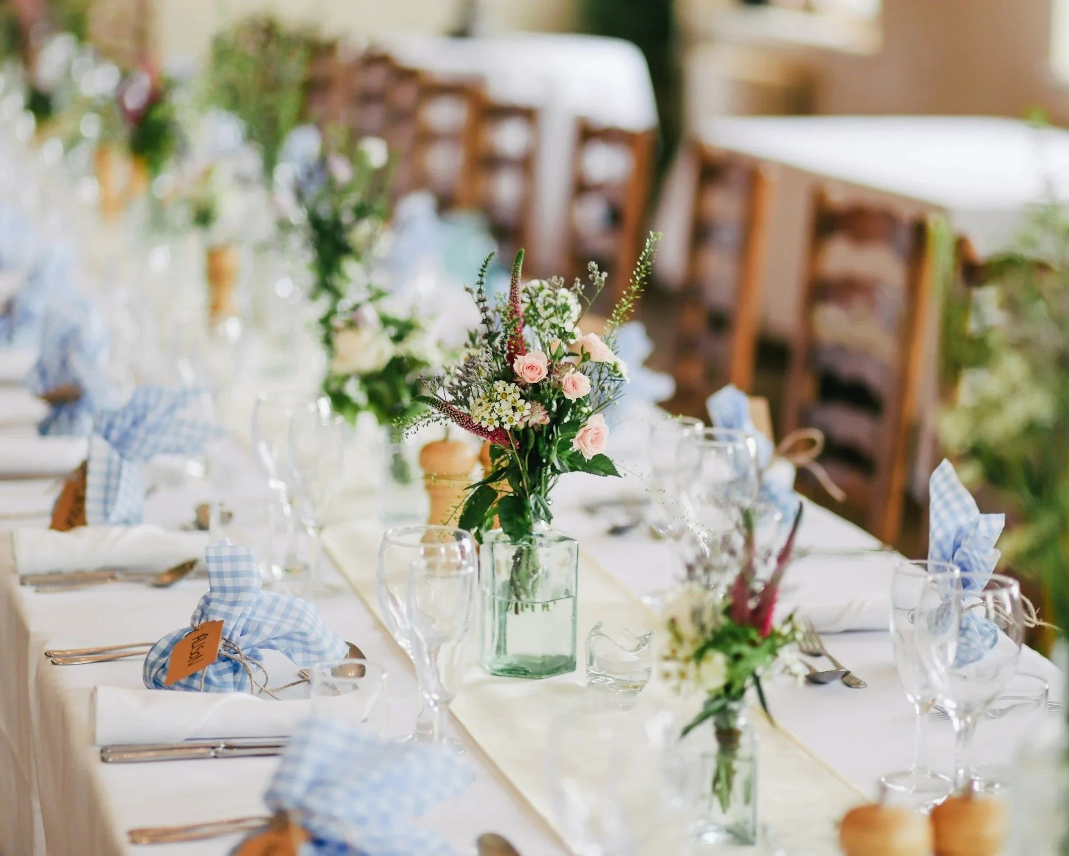 A table set for a formal event, decorated with a floral centerpiece of pink roses and greenery in a glass vase, with wine glasses, blue checked napkins, and silverware arranged around the table. Chairs are visible in the background.