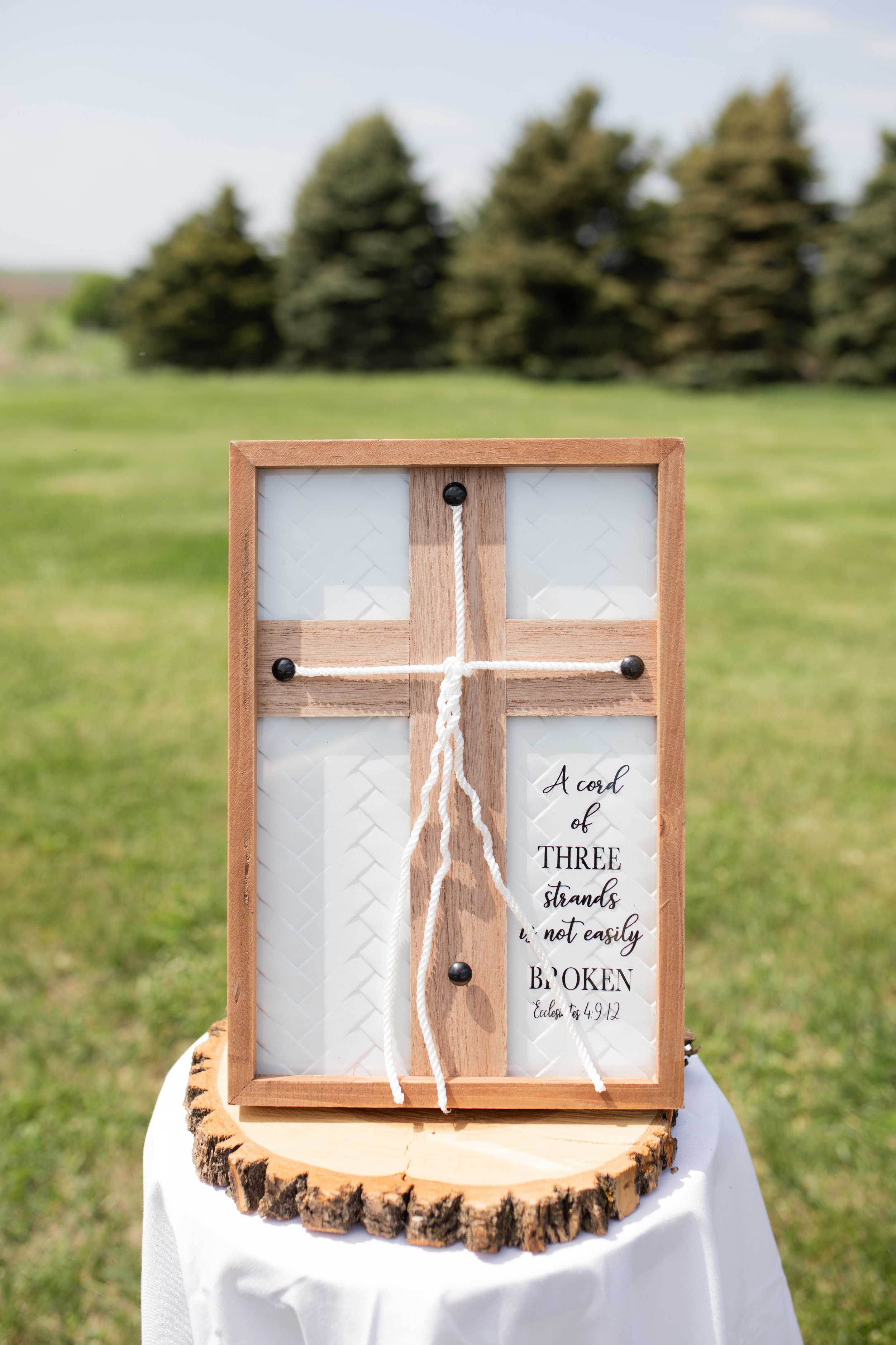 A wooden framed sign with twine, placed on a tree trunk slab, outdoors with blurred trees and grass in the background.