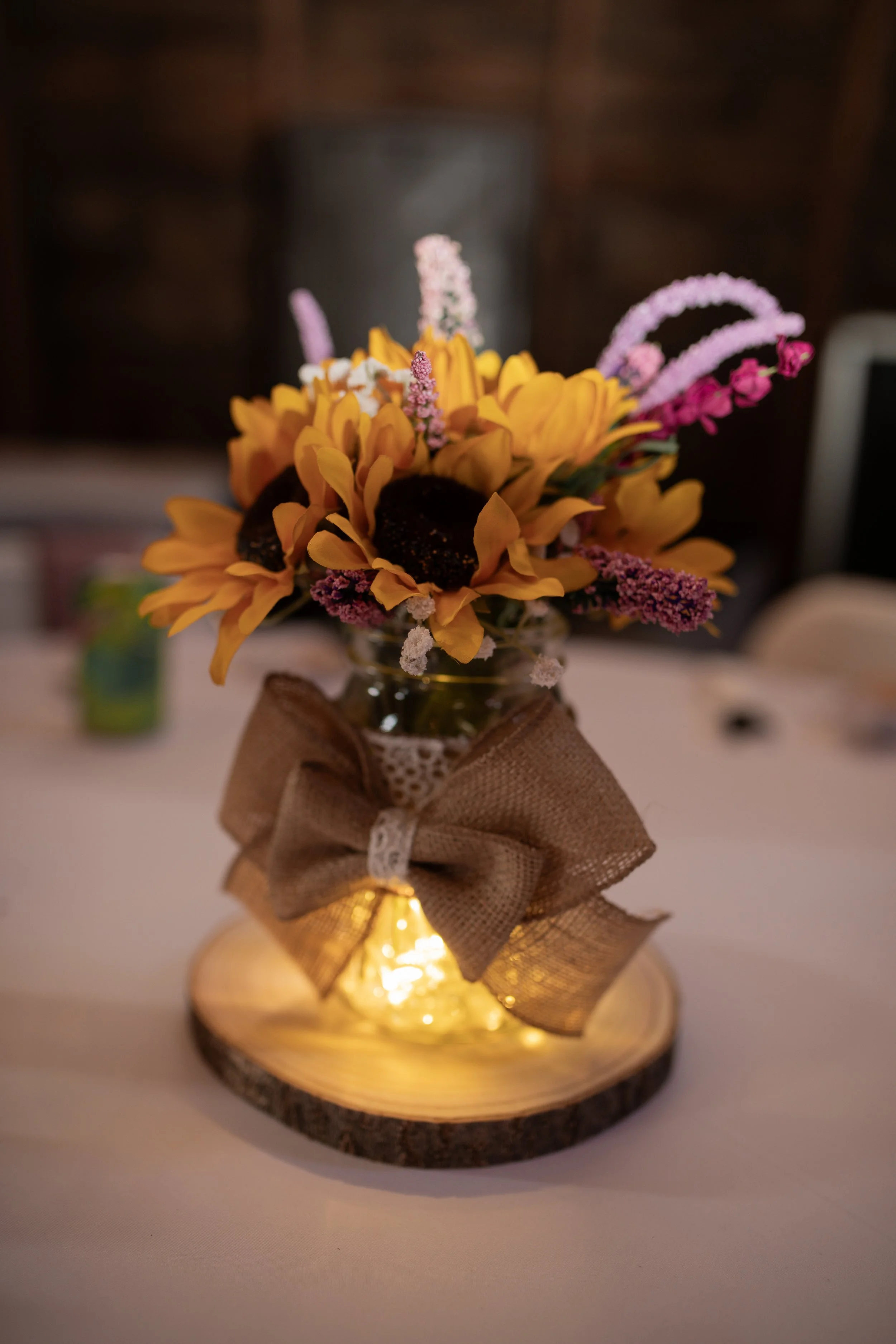 A small bouquet of yellow sunflowers and pink and purple flowers in a glass jar wrapped with a brown ribbon and lace, sitting on a wooden coaster with warm lighting.