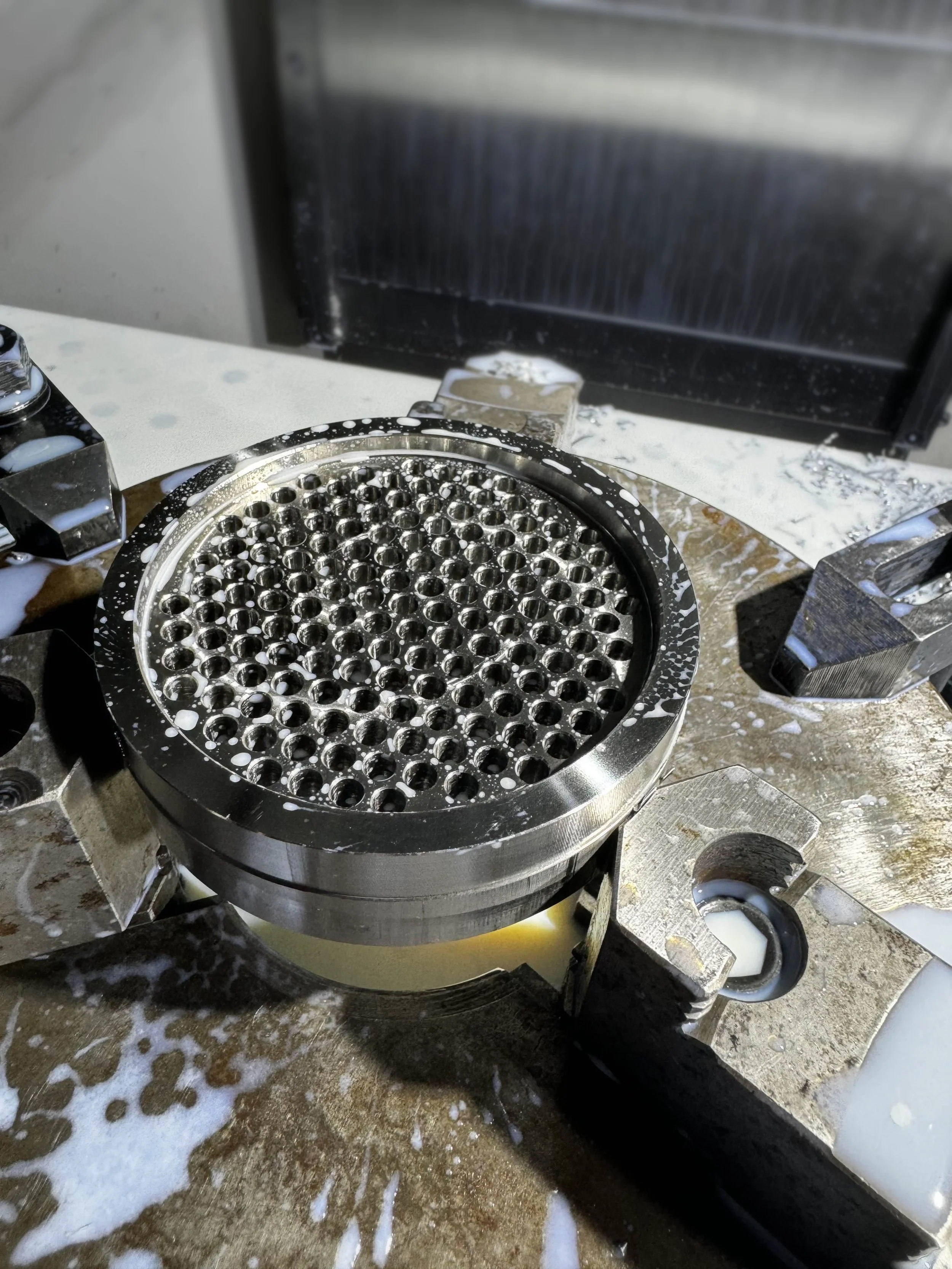 A metal CNC-machined coaster with a perforated plate, surrounded by spilled white liquid and foam on a work surface.