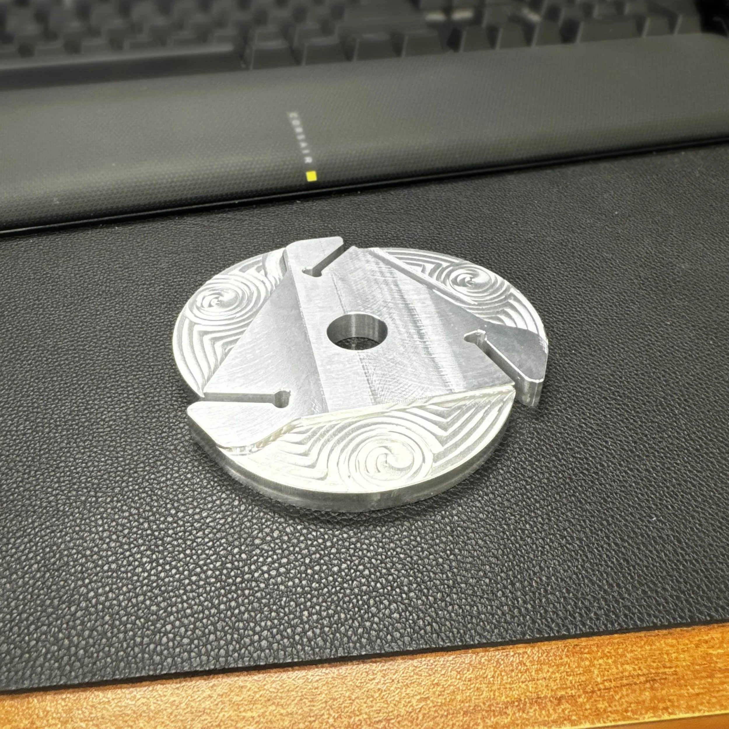 A circular metal component with a central hole, with engraved swirling patterns and three cut-out sections, placed on a dark textured surface with a keyboard visible in the background.