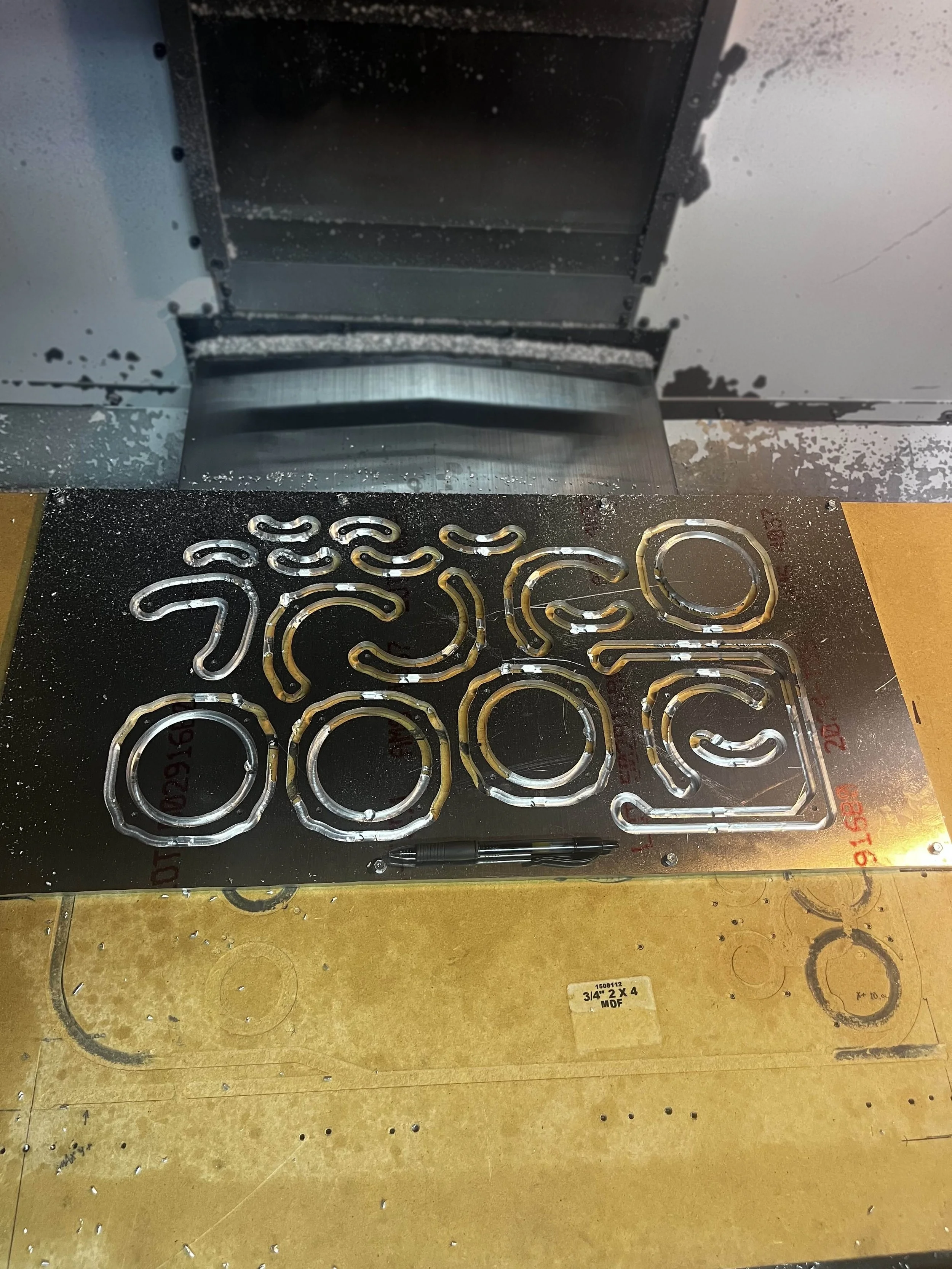 Metal cutouts on a metallic sheet, with a pen placed in front, in a workshop environment.