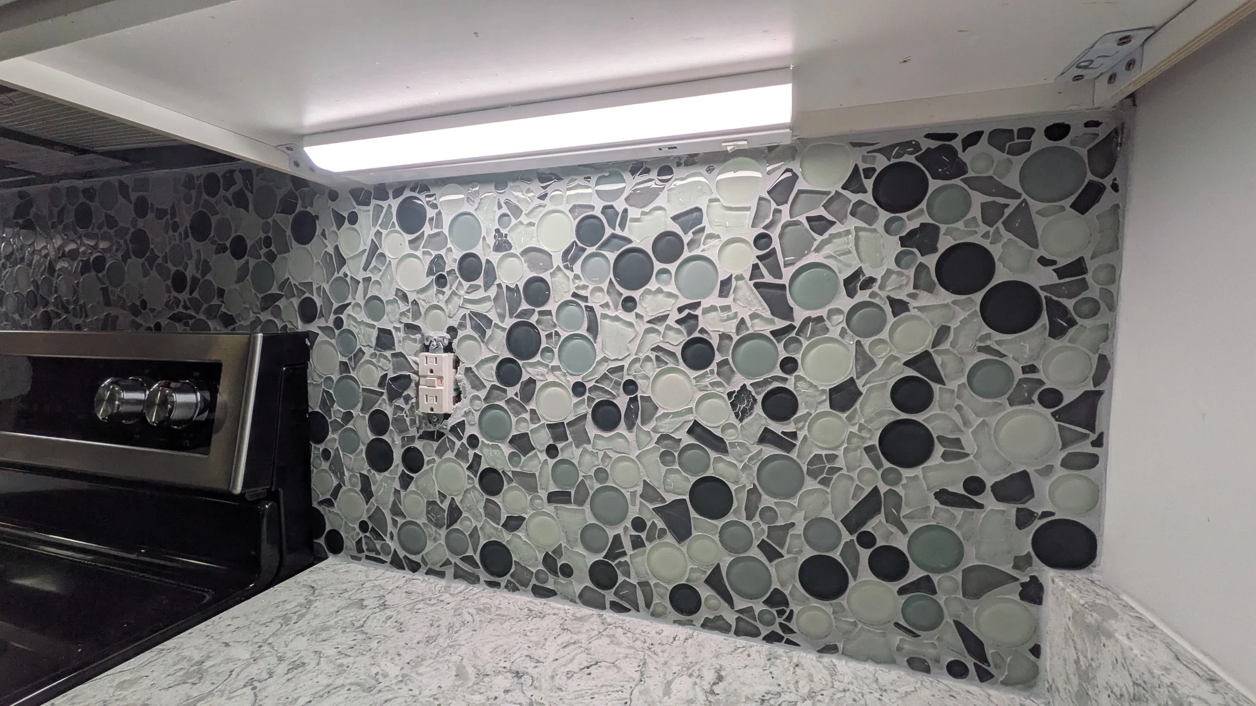 Kitchen wall decorated with a mosaic of various-sized black, white, gray, and clear circular and irregular-shaped glass tiles. Part of a black stove and a marble countertop are visible.